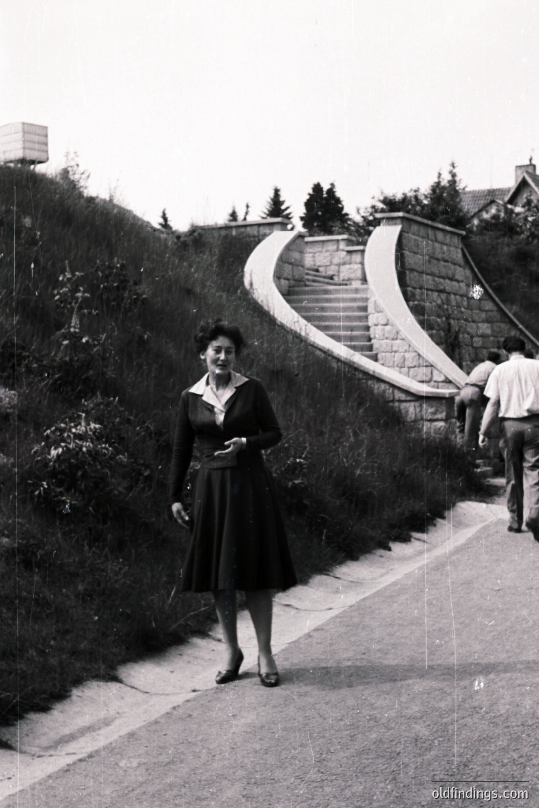 A well-dressed woman stands on a roadside alongside a large stone staircase leading up a hillside. The architecture suggests a resort town or planned community. Likely 1950s-1960s, the style of dress and photographic quality are consistent with this era. Two men are visible in the background ascending the stairs.