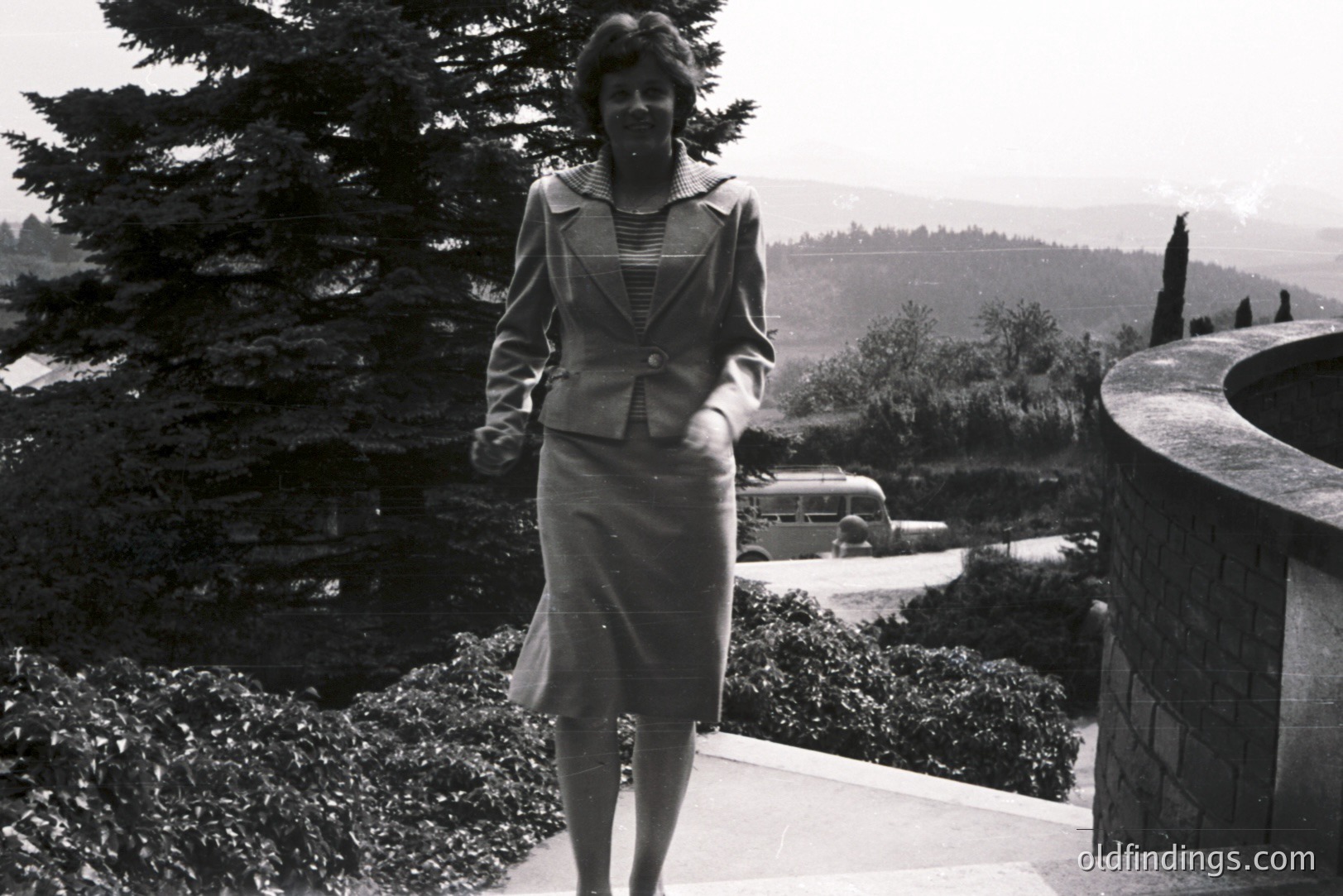 Elegant woman in a tailored suit descends stone steps, overlooking a sprawling landscape with a vintage bus visible below. Likely 1970s, possibly European setting. The architectural details & fashion suggest a design reference. Excellent stock potential.