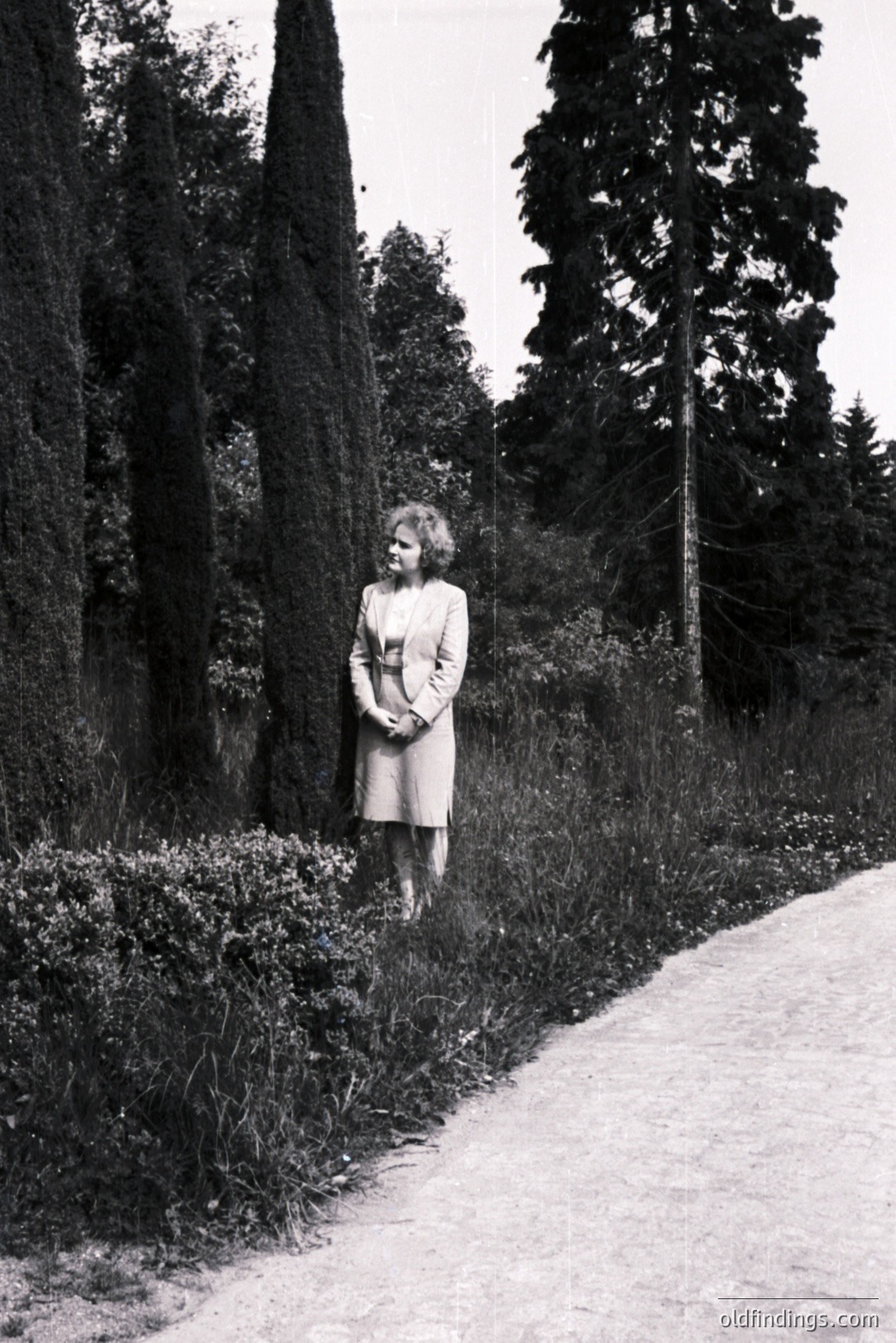 A woman in a knee-length dress and jacket stands beside a gravel path and tall, neatly-trimmed cypress trees. The setting appears to be a formal garden or park with dense foliage and a tall conifer. Likely a mid-century portrait, demonstrating classic styling. Could be useful for vintage fashion or landscape design references.