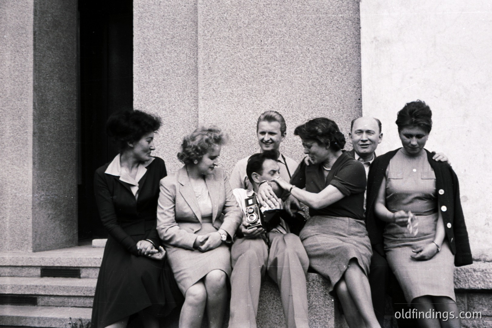 Six individuals are seated on wide stone steps; a man center holds a vintage camera, playfully interacting with a woman who rests her hand on his shoulder. Elegant 1960s attire, including pencil skirts and structured dresses. Possible architectural backdrop suggests a European location. Candid moment, likely a staged group portrait.