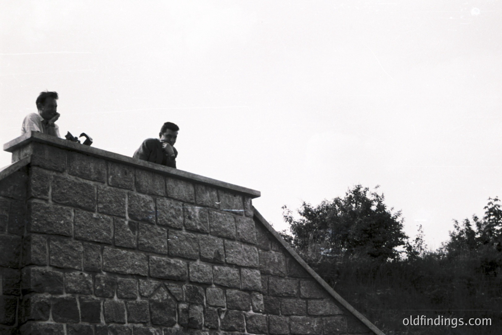Two men in 1960s attire stand atop a stone retaining wall, seemingly engaged in conversation or observation. The wall leads up to a hillside covered in dense foliage. A low-angle perspective emphasizes the structure’s height. Likely taken during a travel or documentary project.