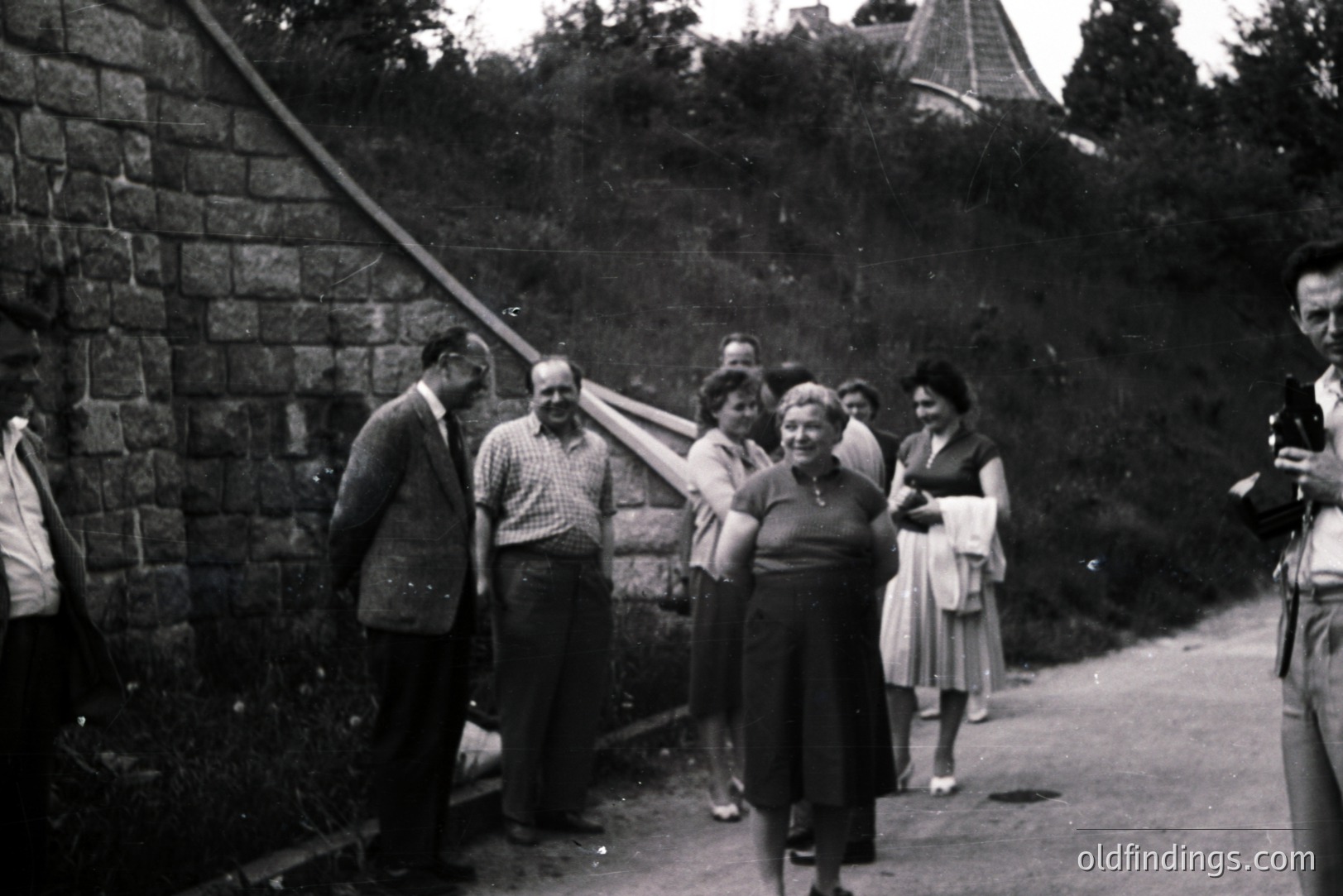 Group portrait; men in suits and casual attire converse with women in 1950s dresses near a stone retaining wall and hillside. A photographer stands to the right, documenting the scene. Likely a family or travel group. Architectural details suggest a European location.