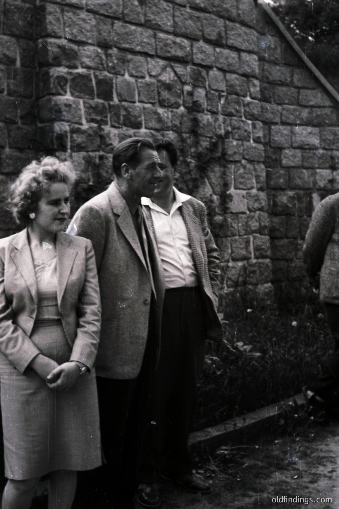 A formally dressed woman and man stand facing away from the camera, leaning against a textured stone wall. The woman wears a tailored suit; the man a tweed jacket over a collared shirt. Likely 1950s, posed studio portrait. Background suggests garden or grounds. Potential stock photography reference.