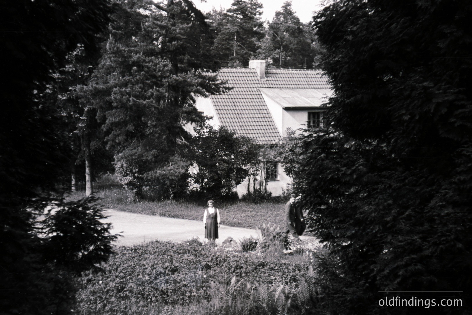 Framed by foliage, a charming, tile-roofed house sits within a lush garden. Two figures in long dresses stand on a driveway leading to the home. Likely a family portrait, the style suggests a mid-20th century, possibly 1950s, European setting. Architectural details and dress styles are noteworthy.