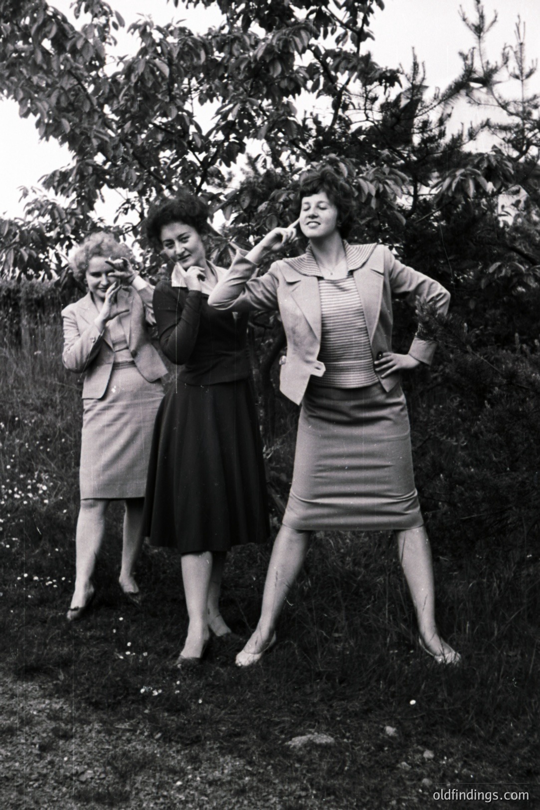 Three women in stylish 1960s suits pose amidst foliage. The group exudes confidence with dramatic stances and visible heels. The textured grass and dense shrubbery backdrop create depth. A snapshot referencing mid-century fashion and group portraiture.