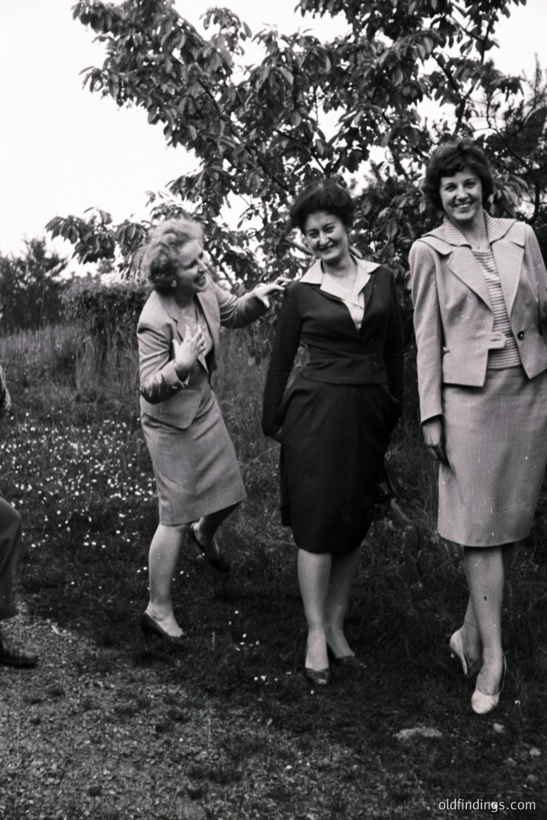 Three women in tailored suits stand outdoors, appearing animated. The background includes foliage and possibly a hillside. A mid-century aesthetic and fashion style are evident, likely 1960s or 1970s. Possible outdoor portrait or candid moment.