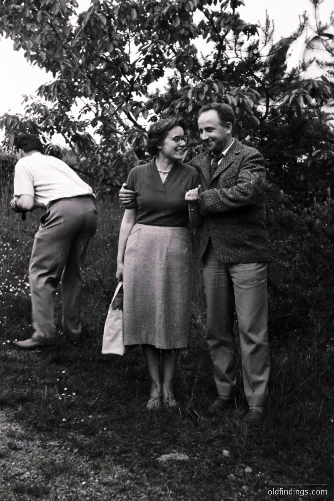 A couple stands in a garden, the man with his arm around the woman; a third figure is partially visible behind them. The scene evokes a mid-century domestic moment, likely 1950s. The woman wears a dark V-neck top and a midi skirt.