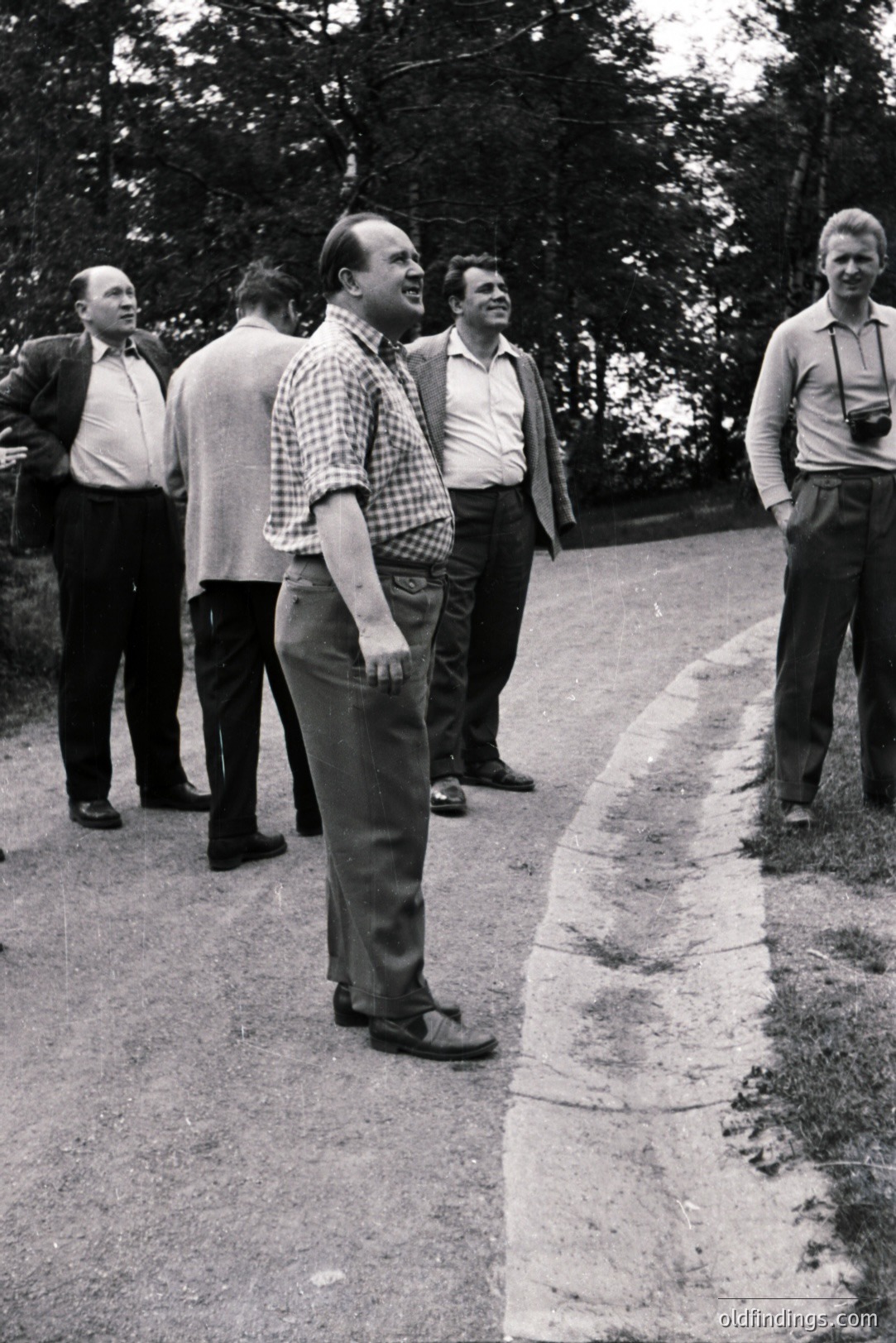 Group of men in suits & casual wear converse on a rural road. The man in the checkered shirt appears to be gesturing. Visible foliage suggests an outdoor, possibly wooded, location. Appears to be a candid snapshot, likely 1970s era. Potential stock photo value.
