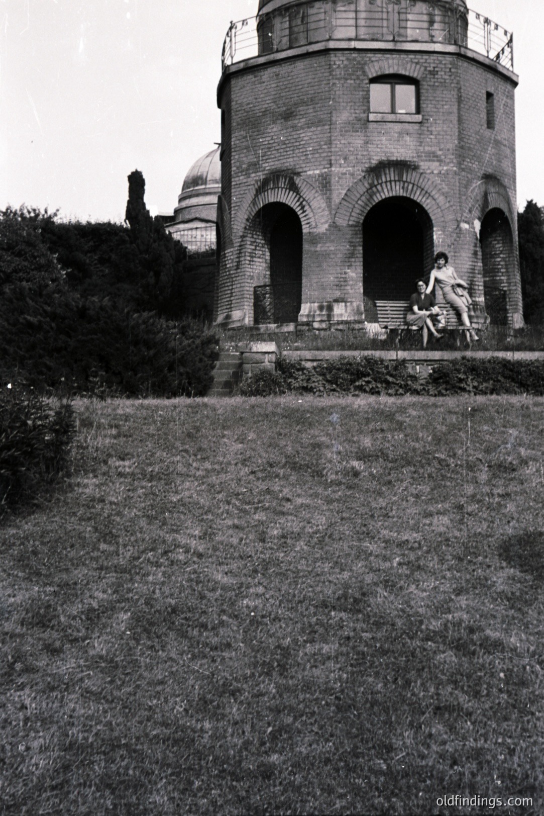 A cylindrical brick structure with arched openings sits atop a slight rise, two figures perched on the base. Dense foliage forms a backdrop; a glimpse of a dome visible behind. Appears to be a park pavilion or folly, possibly 1930s-1950s. Likely a European location. Historic garden design reference.