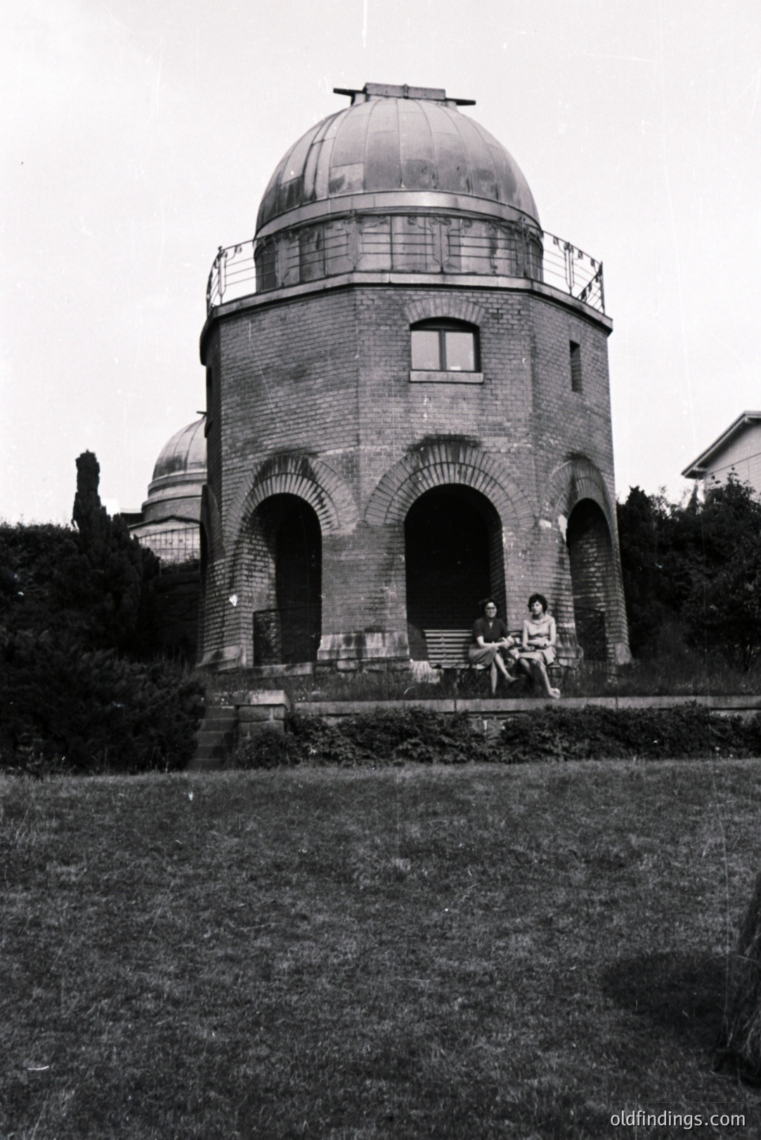 An observatory building with a domed roof sits atop a slight rise. Two women in 1960s attire are seated on a ledge in front. The brick structure features arched windows and a small rectangular window. A second, similar observatory is visible behind.