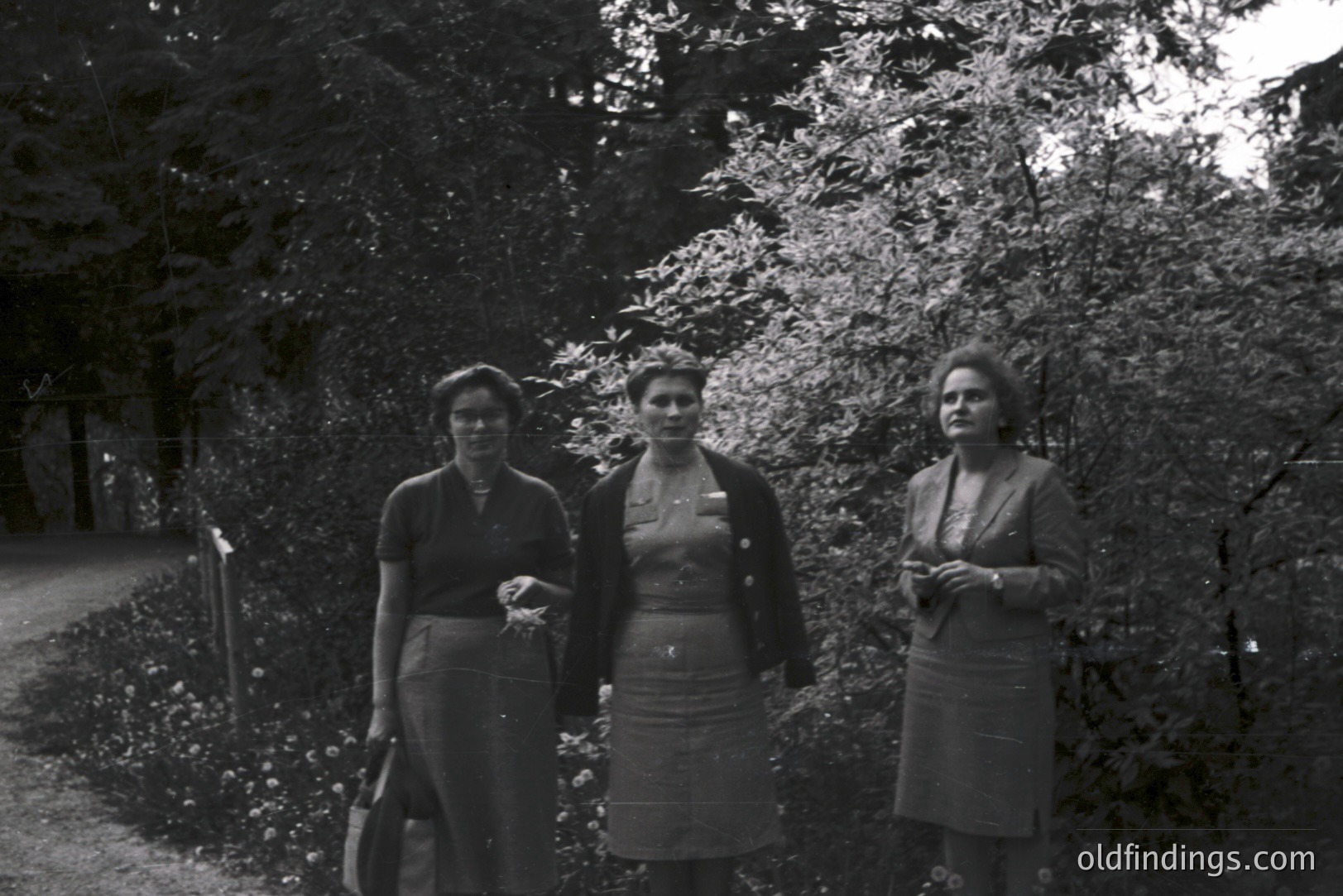 Three women stand in a garden, depicted in a black and white portrait. The composition suggests a candid moment; they are dressed in modest 1950s attire. Lush foliage forms a backdrop. A pathway is visible to the left. Stock photo potential for fashion or mid-century design.