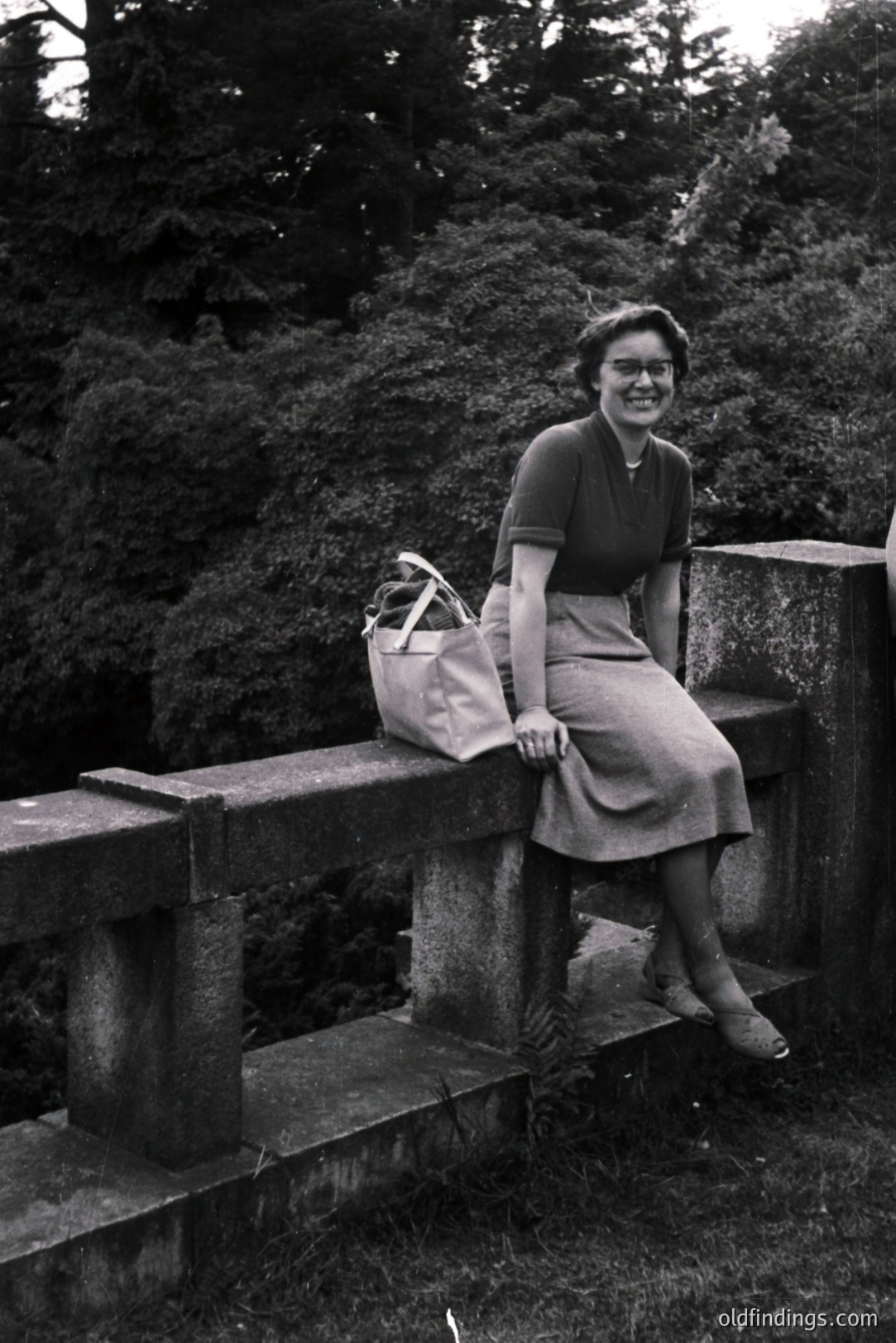 A smiling woman sits on a stone bridge railing, handbag at her side. She wears a fitted dress or top and a midi-length skirt, typical of the 1950s style. Lush greenery forms a natural backdrop. A candid, outdoor portrait. Likely a vacation snapshot.