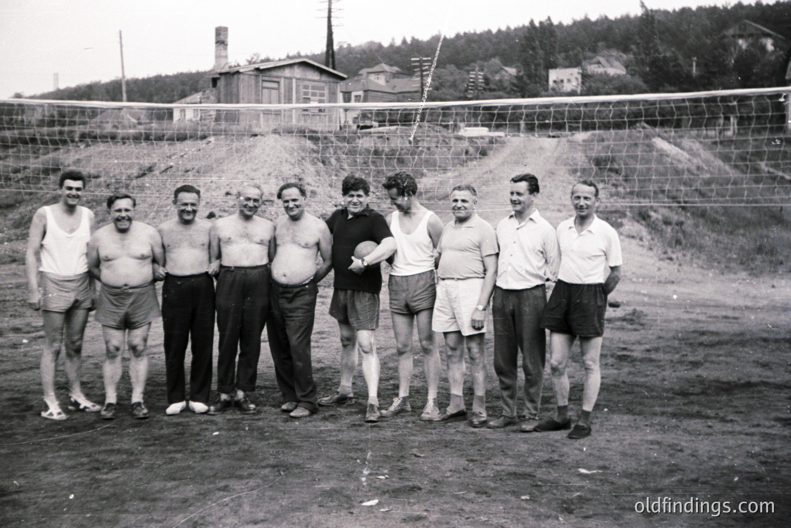 Group portrait of nine men, likely a volleyball team, in a sparse, outdoor setting. They wear athletic shorts and tank tops; one holds a volleyball. Appears to be a rural area with a hillside backdrop & simple structures. Estimate: 1960s - 1970s. Useful for sports history or design reference.