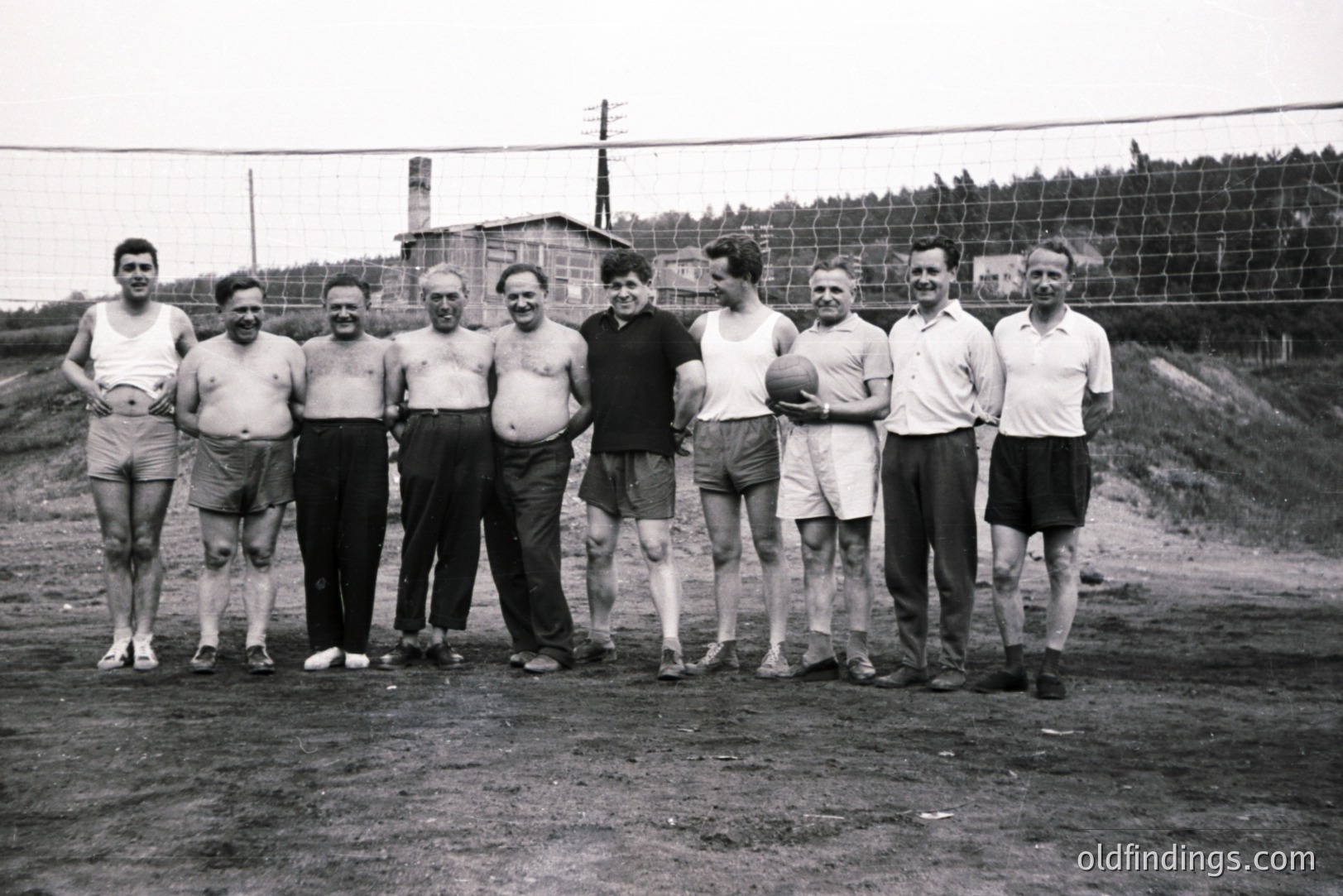 A group of eight men stand beside a volleyball net. Several are shirtless, all wear athletic shorts. A small building with a brick chimney is visible in the background. Likely a recreational or seaside setting. Appears to be 1950s or 1960s.