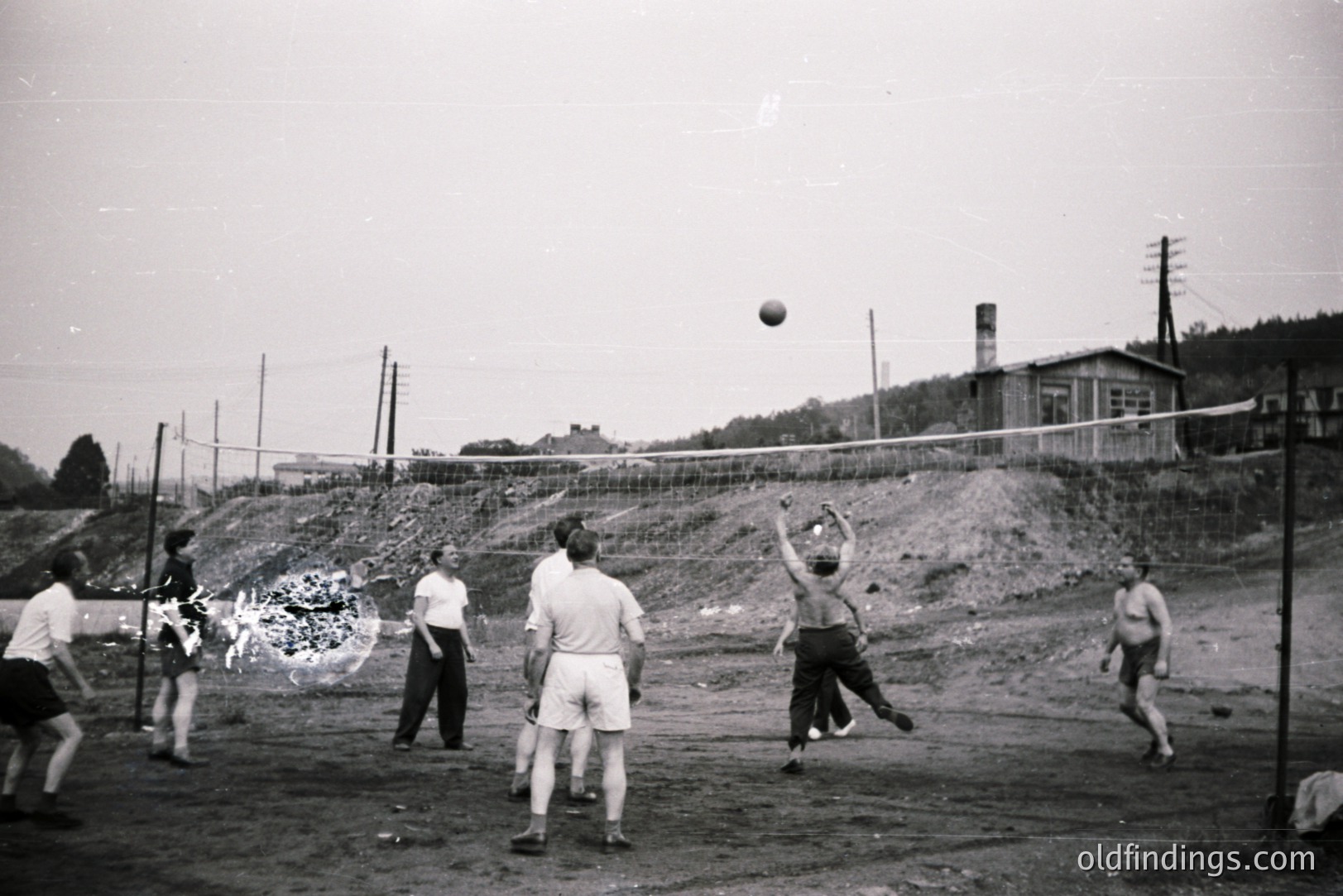 A group plays volleyball on a dirt court. Men in shorts and light-colored shirts compete. A makeshift net divides the space; the backdrop features a chimney, simple buildings, and a hillside. Likely 1950s-1970s, possibly Eastern Europe. Captures recreational activity & post-war infrastructure.