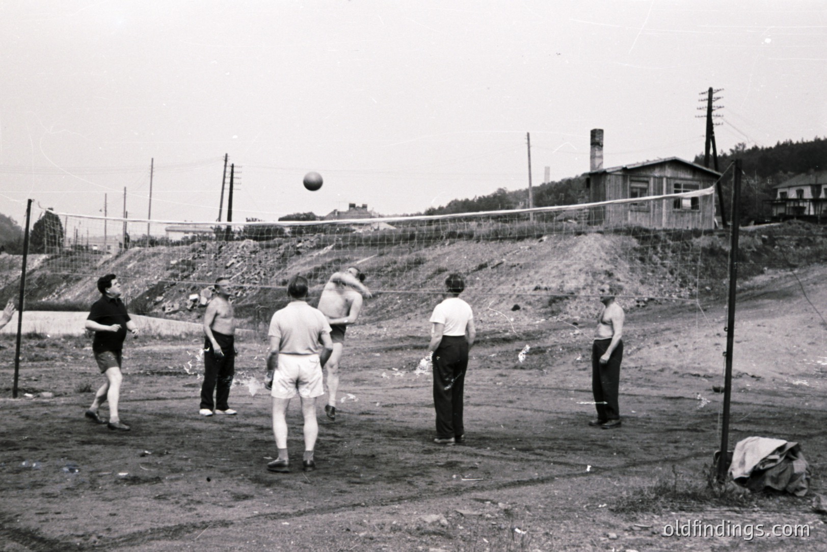 Five men play volleyball on a makeshift court with a wire net. Visible are simple athletic shorts and tops; the background features a hillside with a chimney, power lines, and a rustic building. Likely a recreation area, possibly in Eastern Europe.