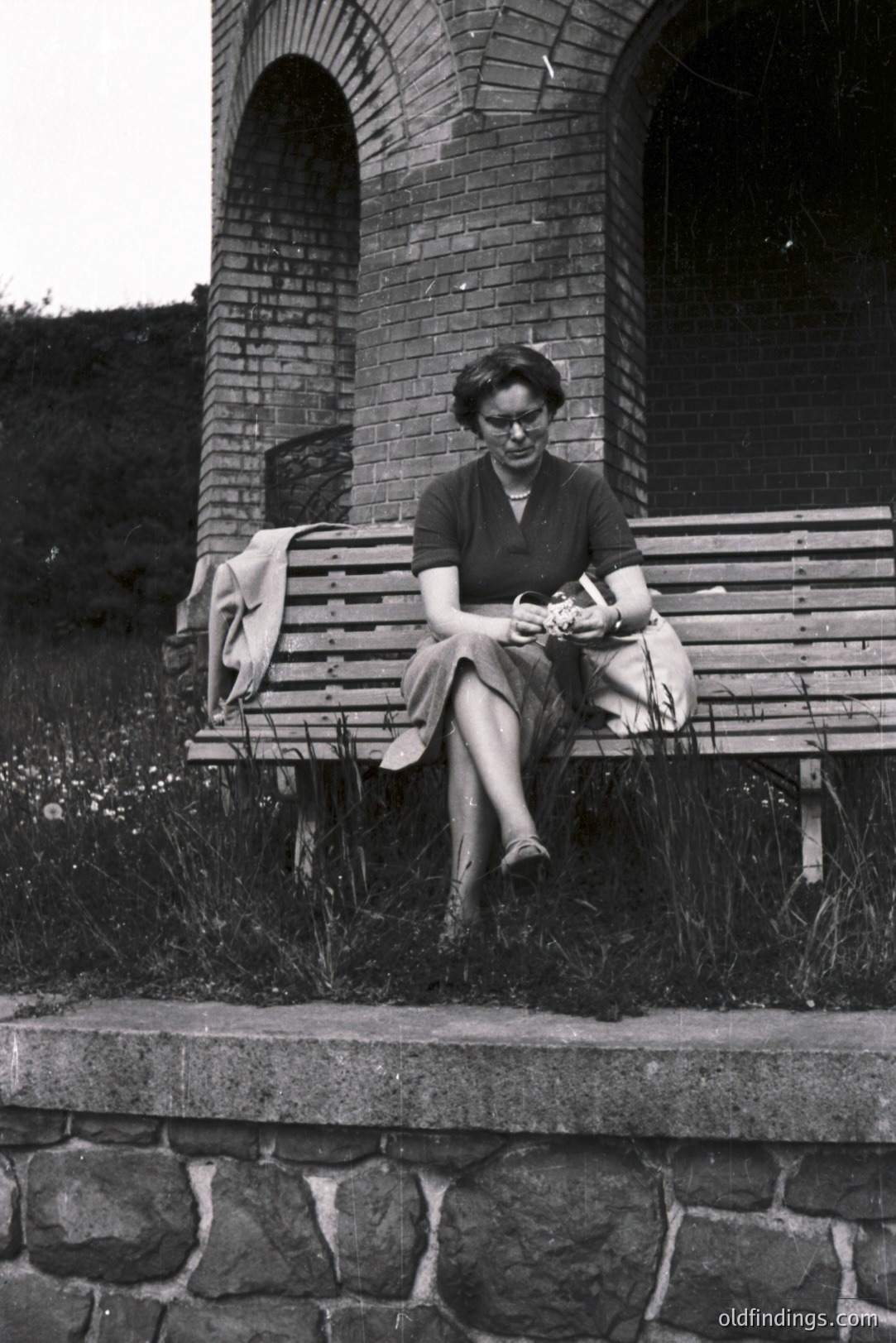A woman in a dark skirt suit and glasses sits on a wooden bench beneath an arched brick structure. She holds a document or papers, appearing contemplative. The setting suggests a public garden or park, possibly European. Likely 1950s-1970s due to the style. Stock potential for vintage portraiture.