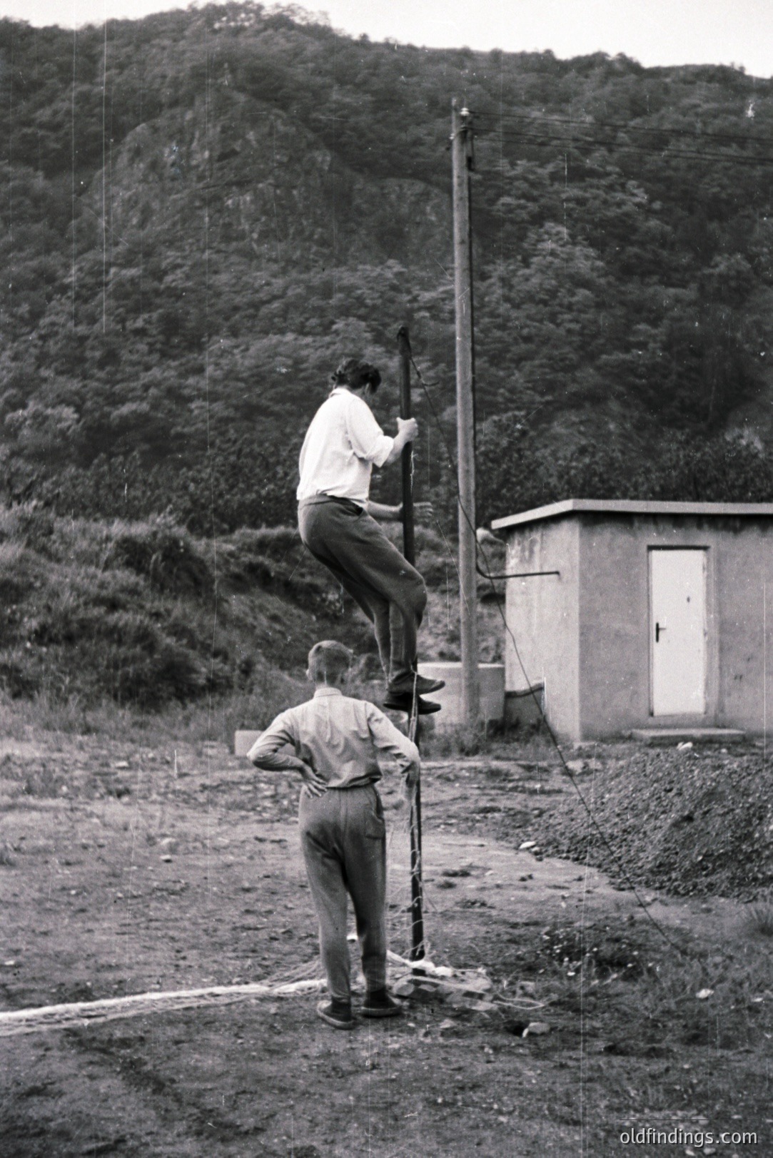 Two young men engage in a climbing game around a utility pole and small concrete building. One is ascending, the other observes. Likely a rural setting with a wooded hillside backdrop. Clothing style suggests a mid-20th century timeframe, perhaps 1950s-1970s. A candid, documentary-style moment.