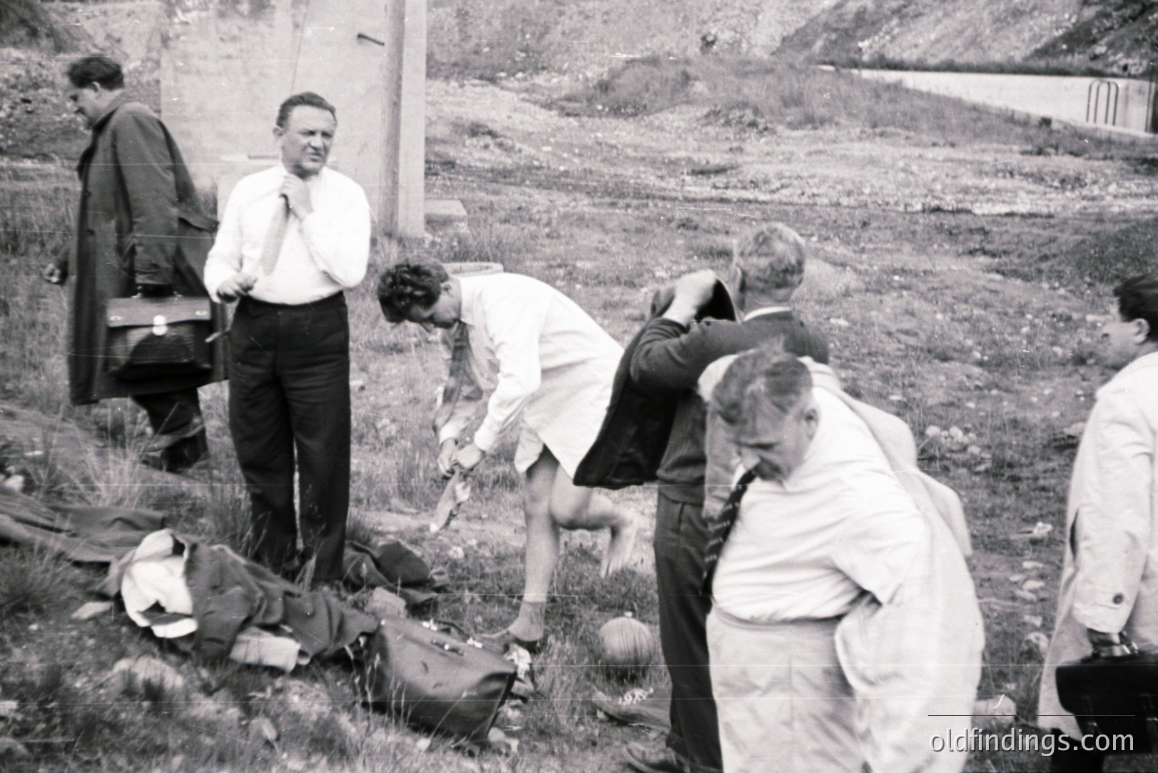 Group of men in suits and lab coats examine a body covered with a cloth on grassy terrain. Appears to be a forensic investigation scene. Likely post-war era, mid-20th century based on clothing and photographic style. Location undetermined, possibly outdoors near a structure.