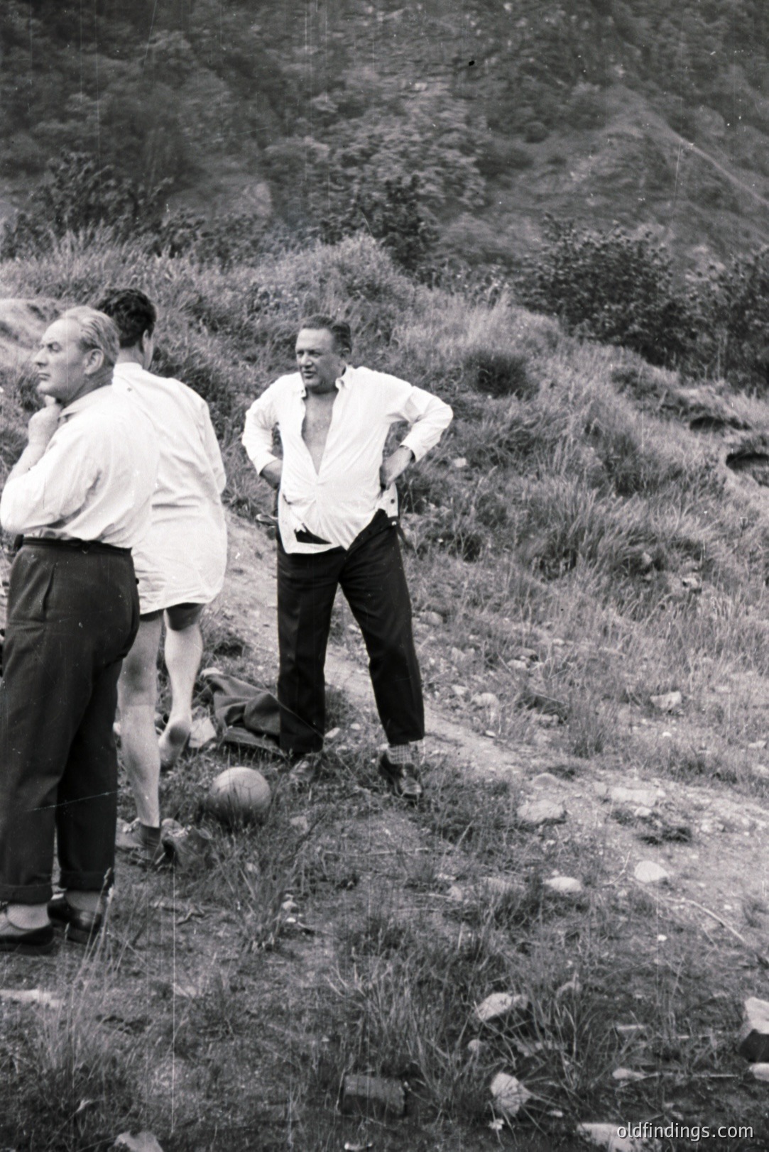 B&W image: Two men in dress pants & open-necked shirts stand on a grassy slope with a small hill/mountain backdrop. One man is barefoot, partially obscured. A tennis ball lies nearby. Likely a candid, informal portrait, possibly 1950s-1970s.
