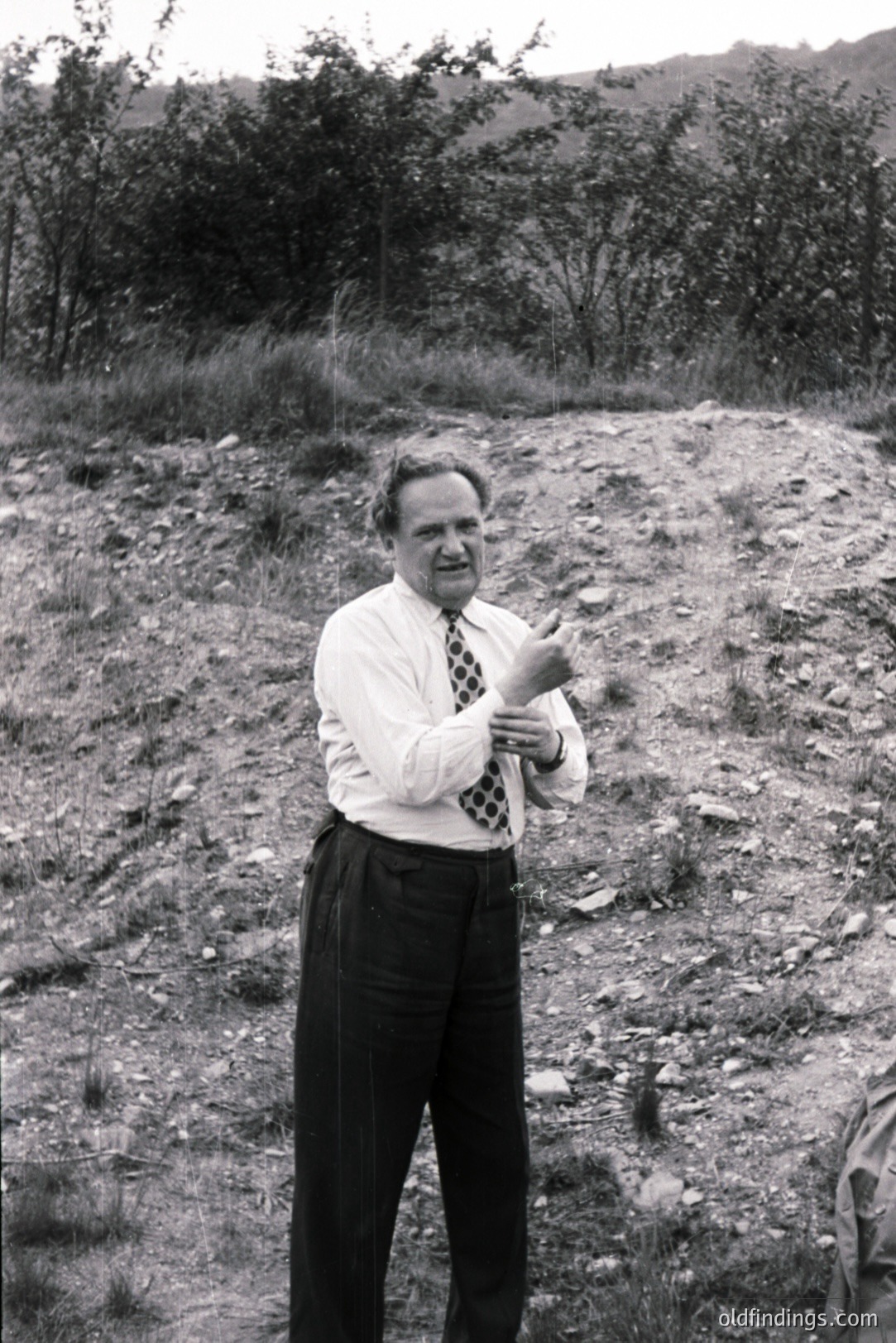 A full-length, black and white photograph depicts a man in a tailored suit and tie, gesturing outdoors on a sloped, grassy area. The man’s pose and attire suggest a professional or academic setting, possibly fieldwork. The backdrop features sparse vegetation and a distant hillside. Likely mid-20th century.
