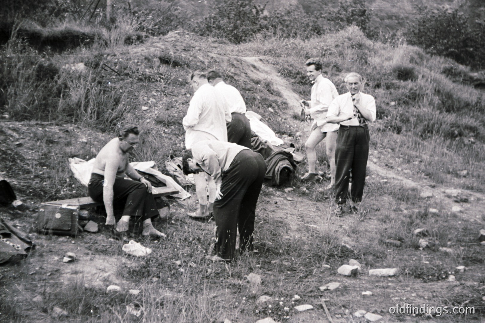 Group of men, dressed formally, appear to be exploring a rocky, overgrown hillside. Several suitcases and bags lie scattered around. One man bends over, examining something on the ground. Appears to be a reconnaissance or survey team, likely 1950s.