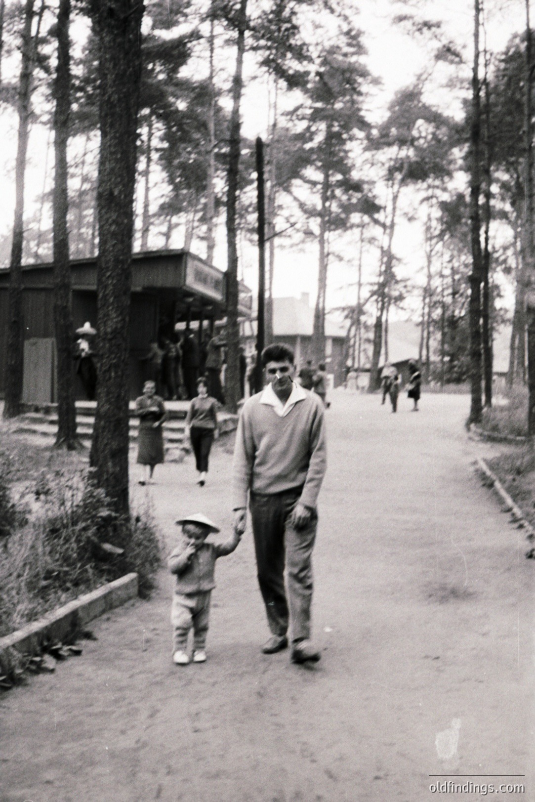 A father and young child walk along a paved path within a wooded area, likely a resort or recreational zone. The child wears a cap and overalls, the man a sweater & trousers. A building and other figures are visible in the background. Appears to be mid-century, possibly 1950s or 60s.