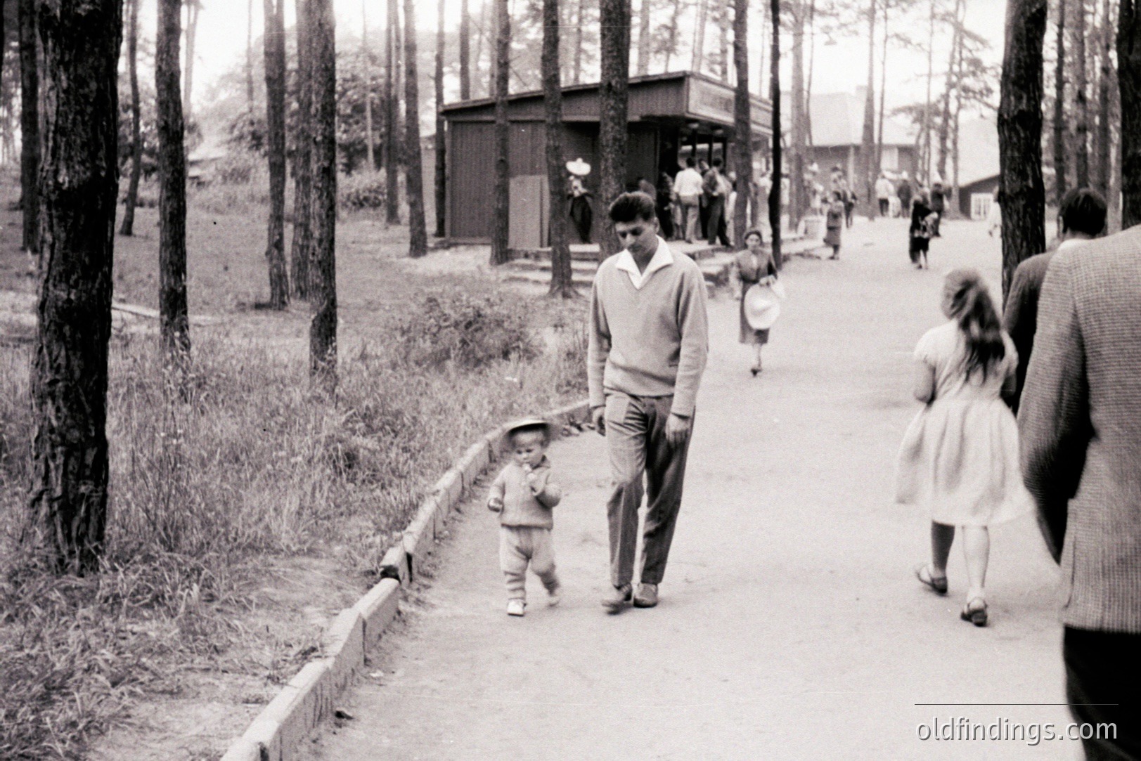 A young boy in a sailor suit walks alongside a man in a sweater, with another young girl following behind. A group of people stand near a building with a peaked roof, visible in the distance. Appears to be a park or recreational area. Likely a family outing, .