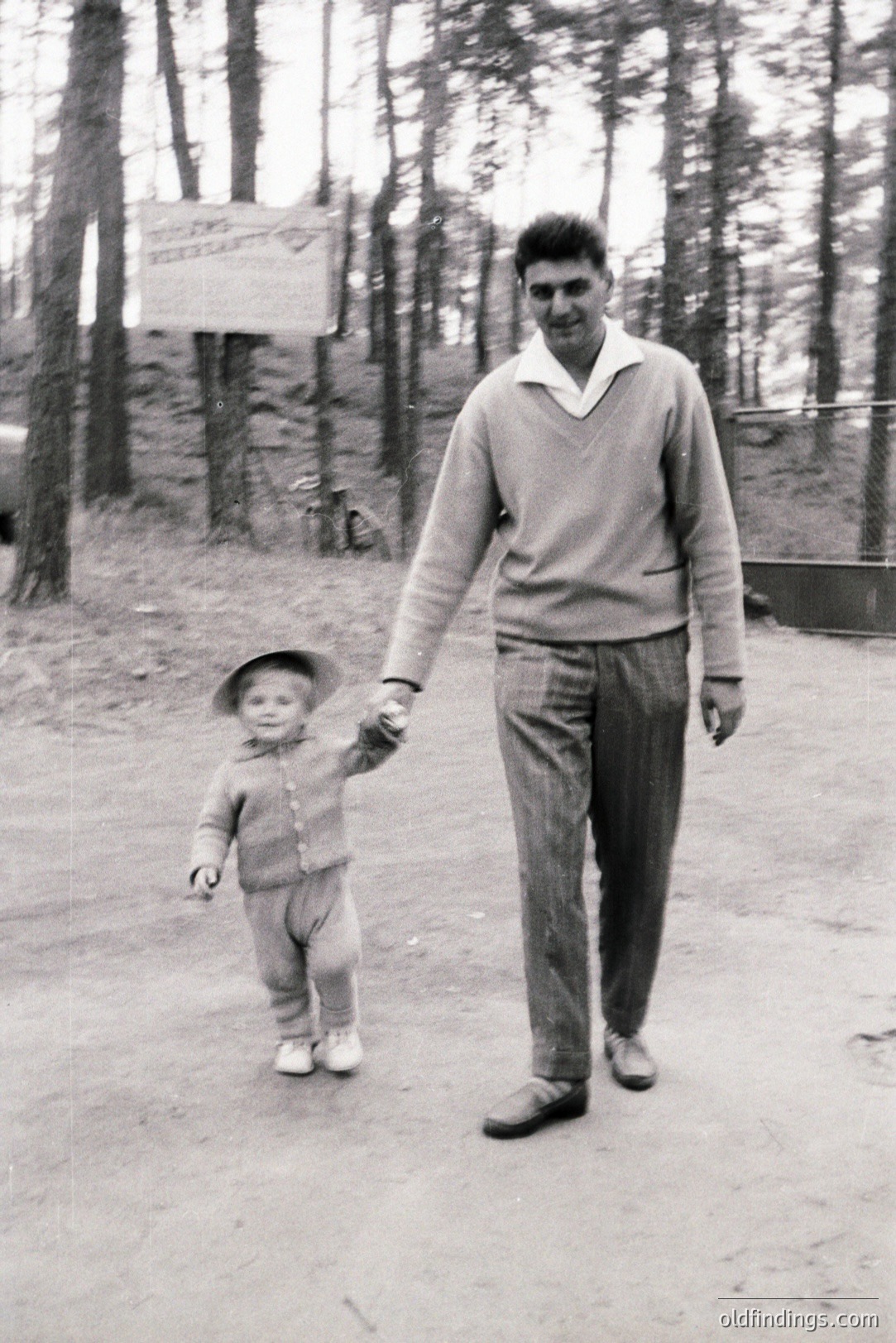 A man and young child walk hand-in-hand on a gravel path, framed by tall pine trees. The man wears a V-neck sweater, collared shirt, and patterned trousers; the child a knit sweater and small hat. Appears to be a casual, candid moment. Likely a family outing. Estimated timeframe: 1960s-1970s.