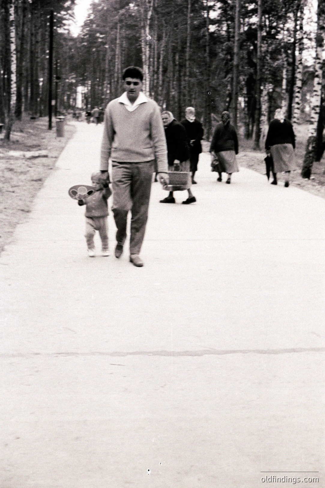 A young man in a cable-knit sweater leads a toddler with a toy in a stroller along a paved path lined with birch trees. Several other figures are visible further down the path. Likely a park scene, c. 1960s-70s. Nostalgic family portrait.