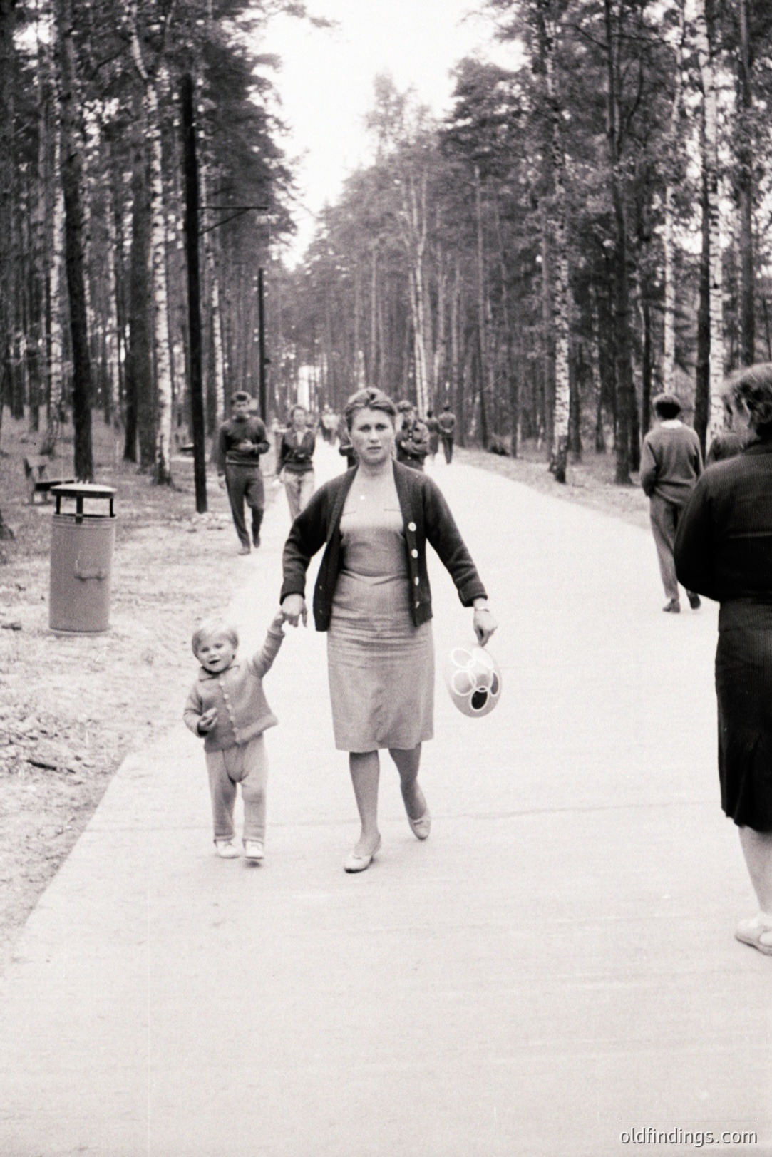 A woman walks along a paved path with a young child, both appearing lively. She wears a cardigan and knee-length skirt, holding a balloon. Background features a birch forest and distant figures. Likely a public park setting, possibly 1960s. Evokes nostalgic family moments.