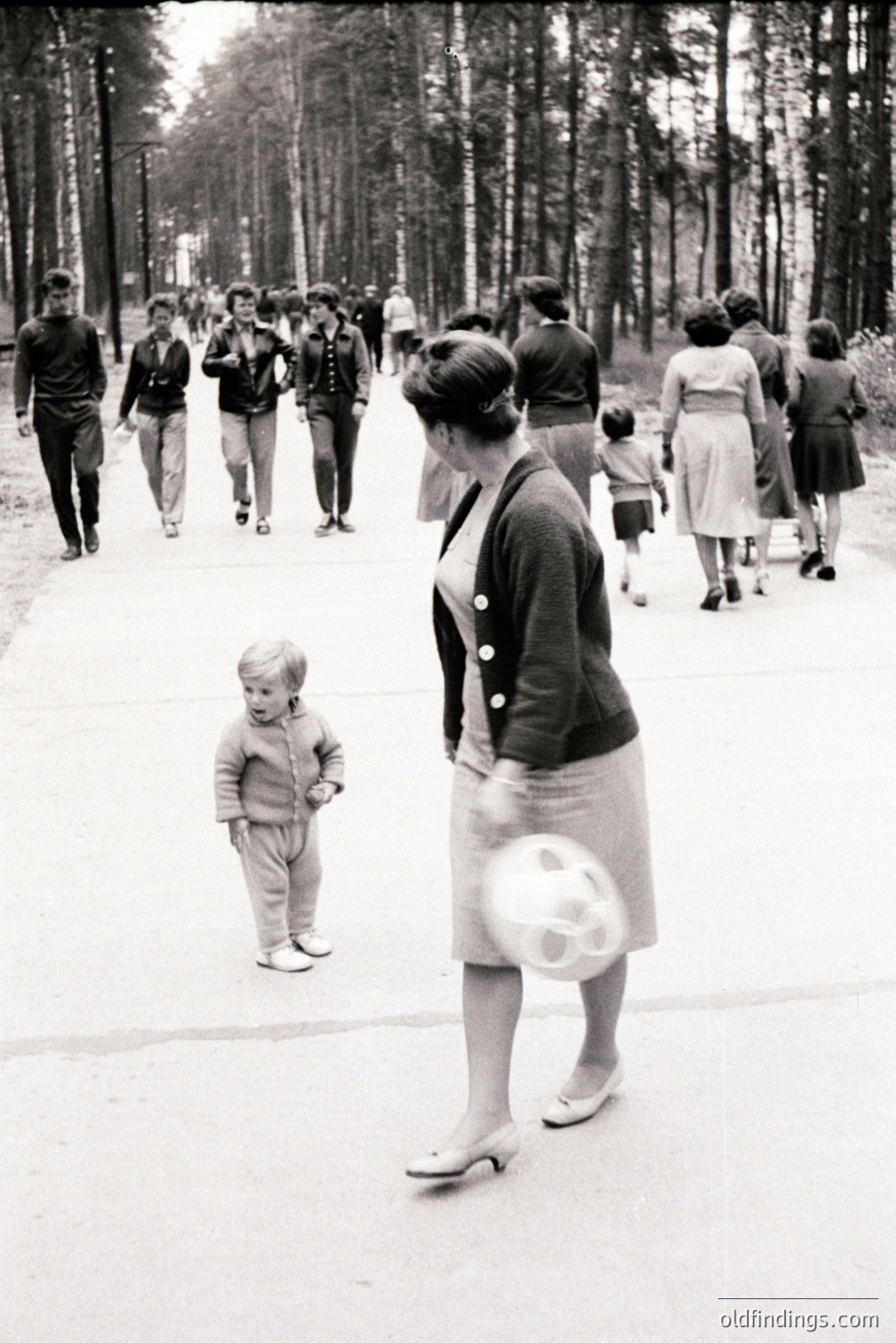 Black and white image shows a woman in a skirt suit & heels walking with a ball, a young boy looks back, amidst a group of people on a tree-lined path. Likely a leisure outing, reflecting 1960s mid-century fashion and family life. Apparent focus on the child’s perspective.
