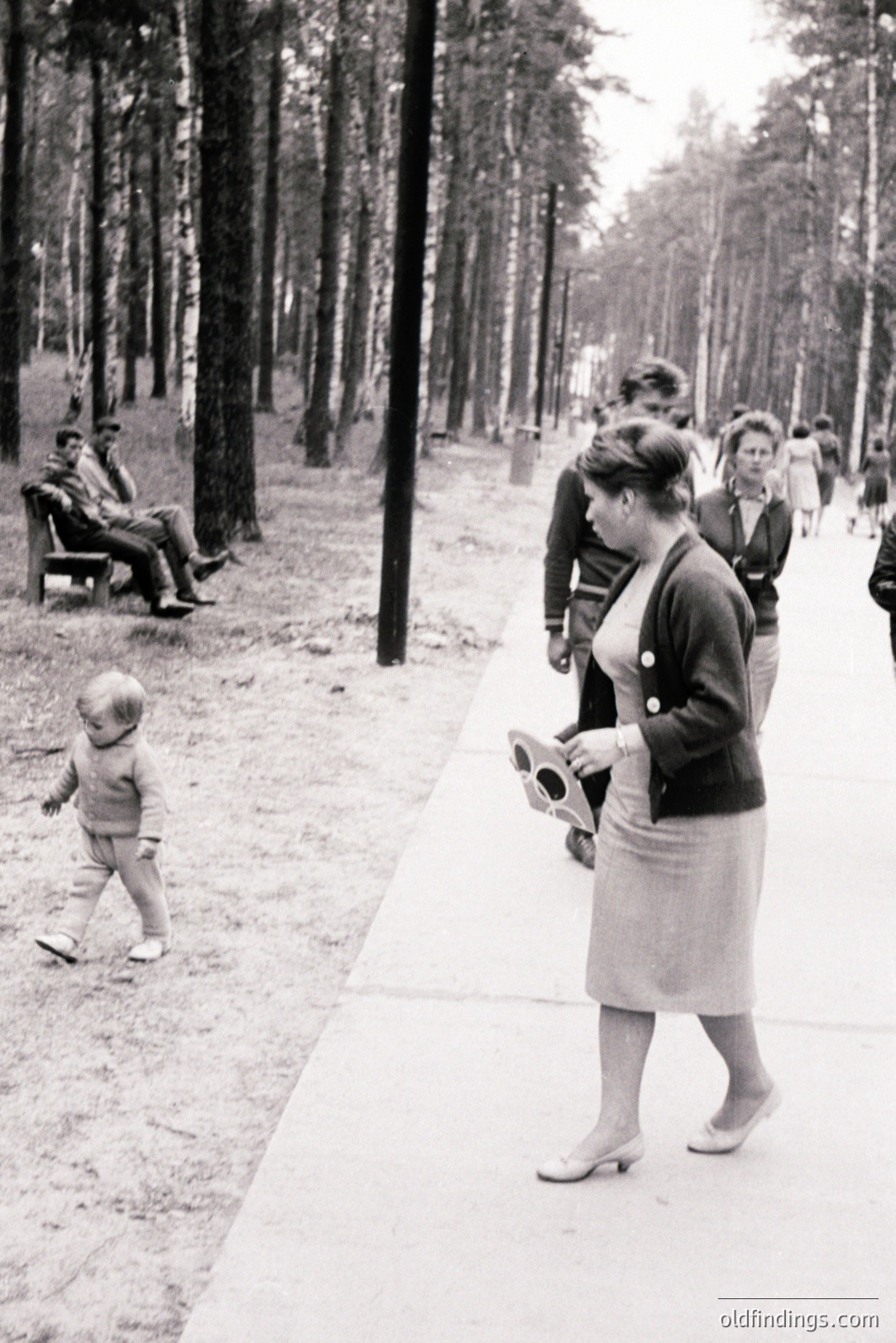 A candid street scene depicts a young boy taking tentative steps, a woman in a fitted skirt suit and heels approaching, and a man seated on a park bench. The setting appears to be a city park with tall trees lining the sidewalk. Likely 1960s, Eastern European style. Evokes nostalgia, family portraits, street photography themes.