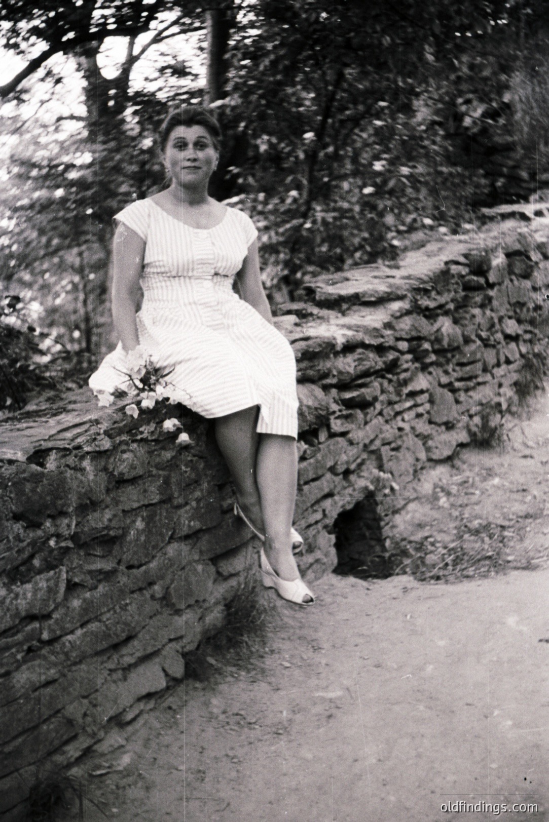 A seated woman in a knee-length, patterned dress and heels poses near a stone wall. She holds a bouquet of flowers. The setting appears to be a garden or park with a paved path visible. Appears to be from the 1960s. Suitable for vintage fashion reference.