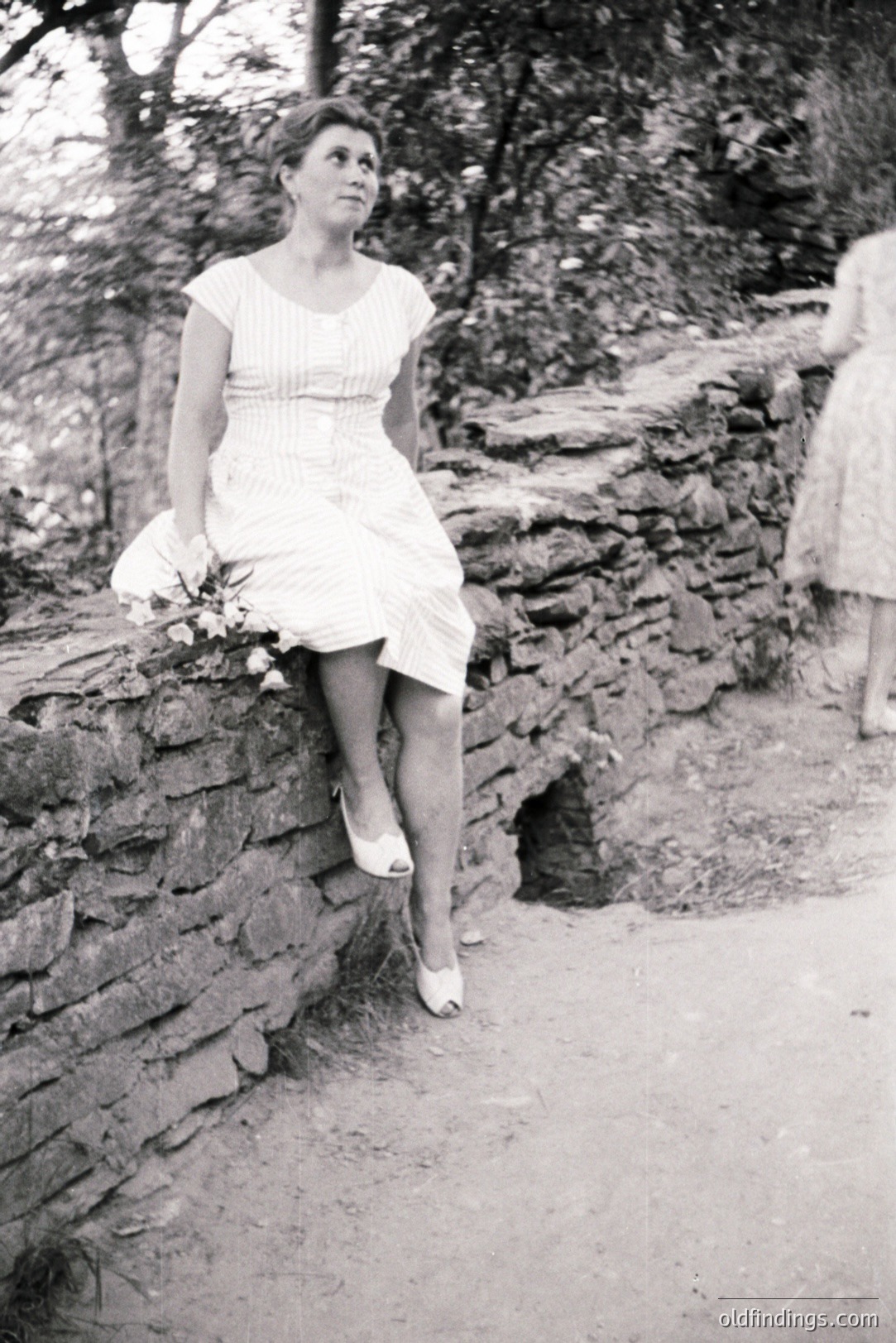 A young woman in a short, patterned dress and heels poses on a stone wall. A second figure, also in a dress, is blurred in the background. Likely taken in the 1960s, the image portrays an informal, candid moment. Could be suitable for design referencing vintage fashion.