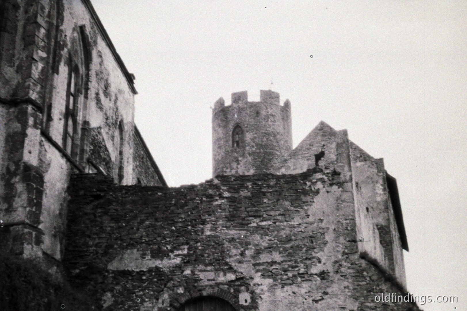 Weathered stone architecture with a crenellated tower rises above a crumbling wall. Textured surfaces suggest age and exposure. Likely a fortification or castle remnant. Possible European location, early to mid-20th century based on photographic style. Valuable for historical context & architectural reference.