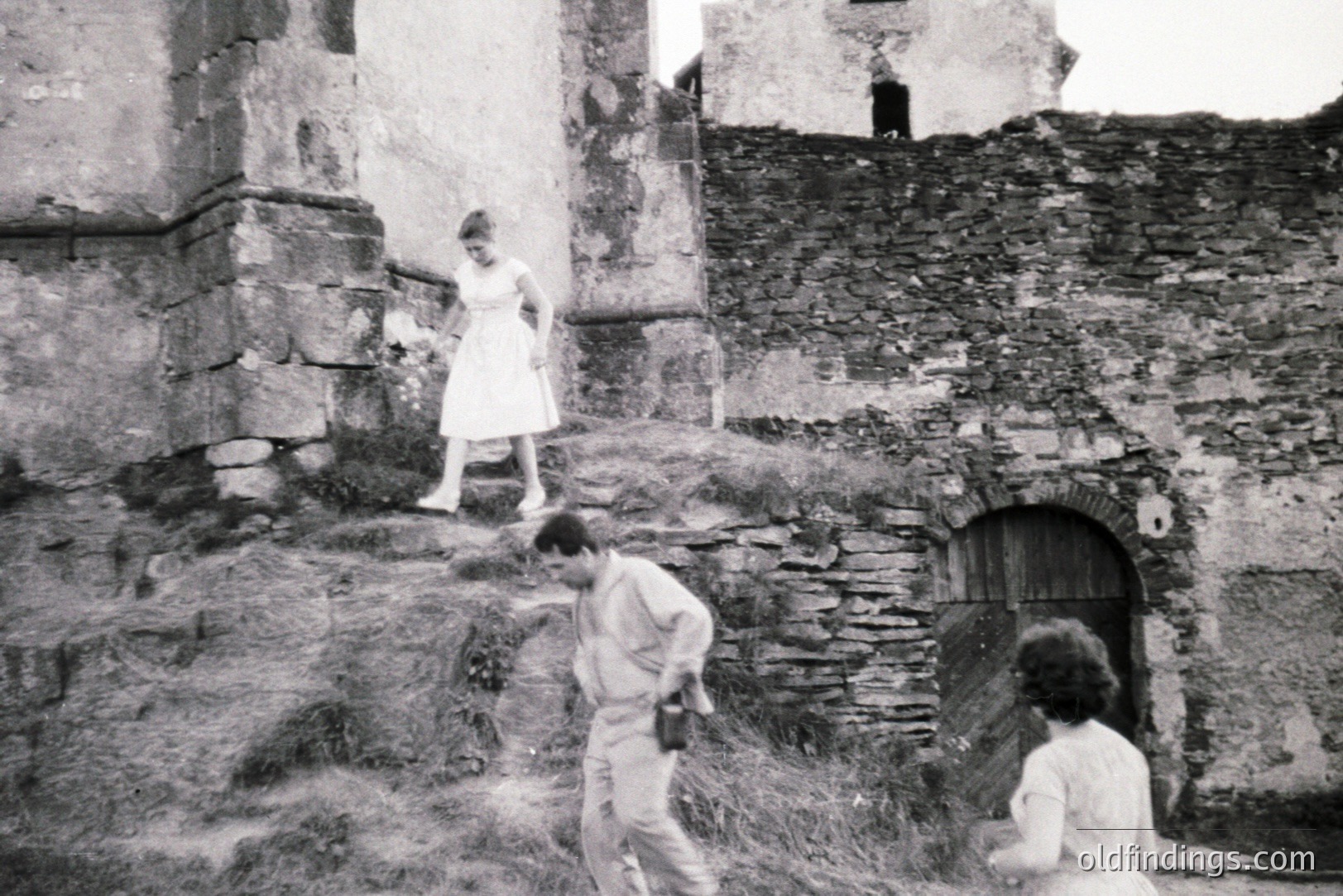 Three figures traverse a rubble-strewn hillside abutting a weathered stone structure with a weathered wooden door. The woman wears a white dress; the men are in light-colored shirts and trousers. Appears to be an informal, candid moment captured in a rural, possibly European setting.
