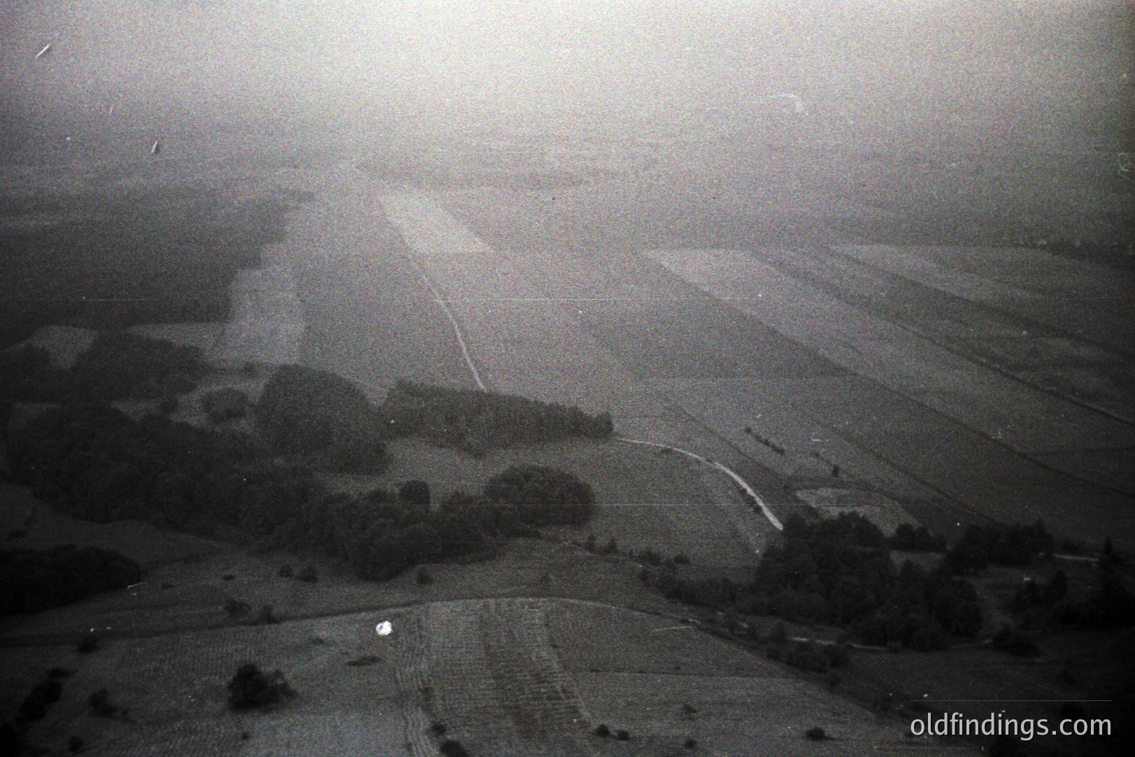 Aerial view showcasing a vast, flat landscape with agricultural fields and scattered tree groupings. A road bisects the scene, leading towards what appears to be an airport or airfield. The monochromatic palette and visible film grain suggest a vintage photograph, potentially from the 1960s-1980s. Likely European, origin undetermined.