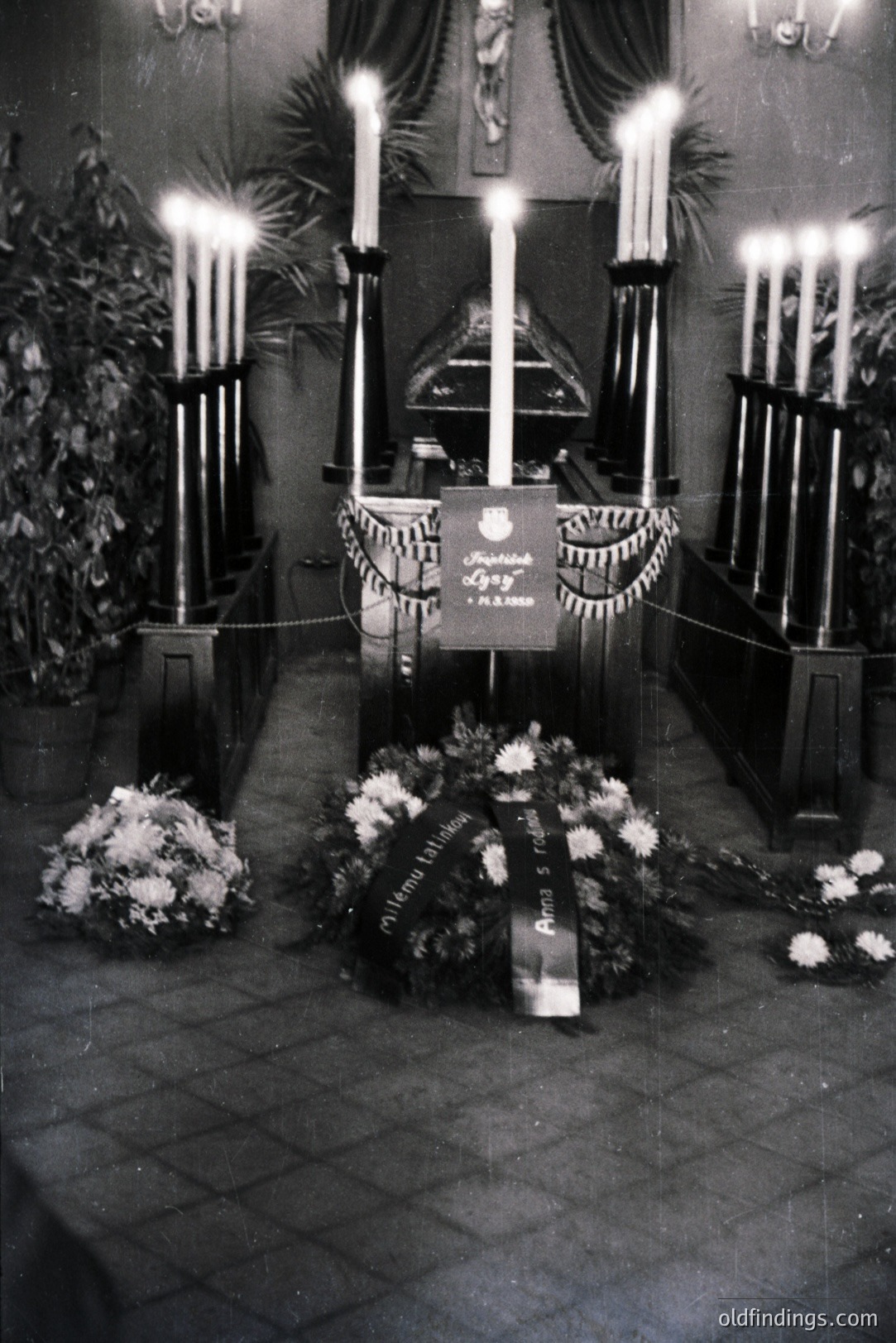 Solemn memorial scene within a church, featuring tall candelabras, floral arrangements, and a displayed plaque bearing text in what appears to be Hungarian. Likely a commemorative event. Visible ropes indicate a restricted area. Appears to be late 1960s/early 1970s.