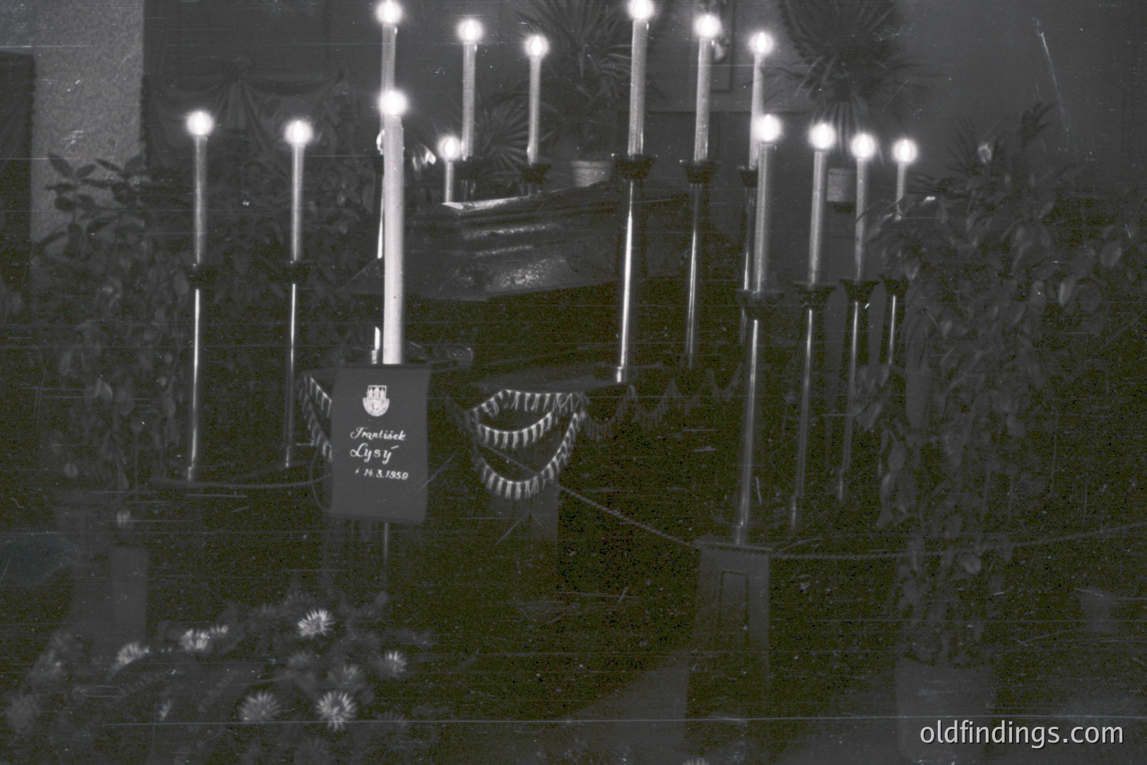 A formal, dimly-lit indoor scene with numerous tall, lit candles arranged around a draped table. A flag or banner with Cyrillic text is visible, likely indicating a memorial or commemorative event. Appears to be a staged, possibly religious or patriotic display.