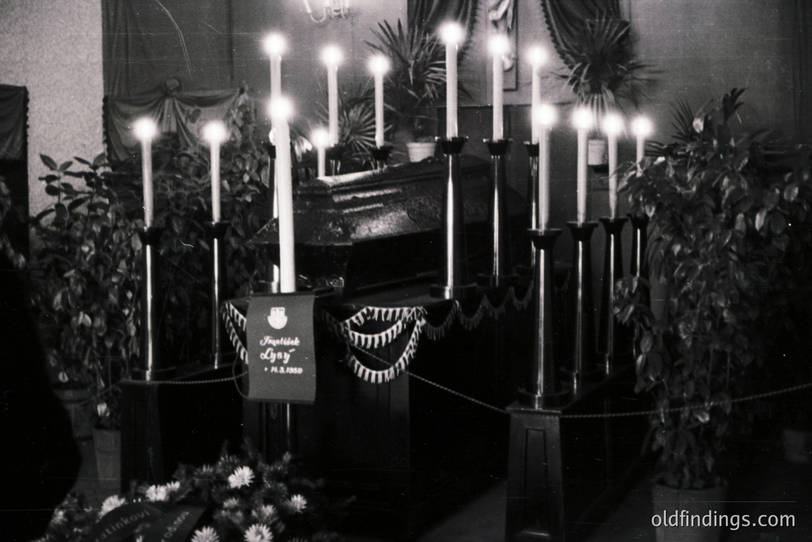 A tall, dark altar display features numerous lit candles, arranged within a multi-tiered structure and framed by potted plants. A decorative banner with Cyrillic script hangs prominently. Likely a religious ceremony or memorial, possibly in a Slavic country. Appears to be a vintage photograph.