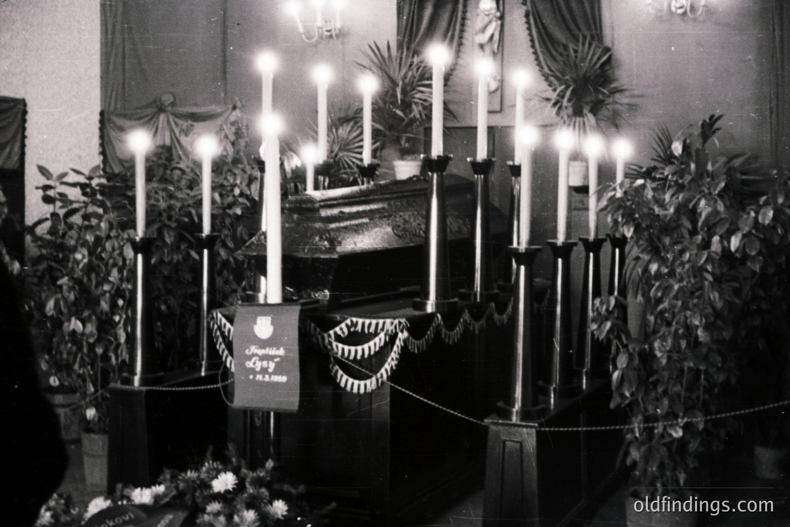 Elegantly draped casket displayed within a room, surrounded by tall, lit candles and potted greenery. A black banner with Cyrillic text and a coat of arms is affixed to the front. Likely a state funeral or memorial service.
