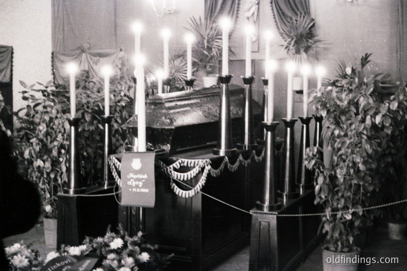 Solemn memorial scene: A casket draped in fabric, encircled by tall, lit candles and potted plants. A banner bears Cyrillic script & a crest. Likely a state funeral or public mourning display, mid-20th century. Stark, formal composition.