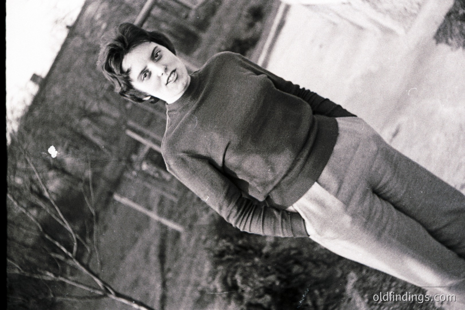 Young woman in a cable-knit sweater and high-waisted trousers stands outdoors, posing with a direct gaze. Architectural details hint at a residential setting, possibly European. The high-angle shot and clothing suggest a 1970s aesthetic. Likely a personal portrait or fashion reference.