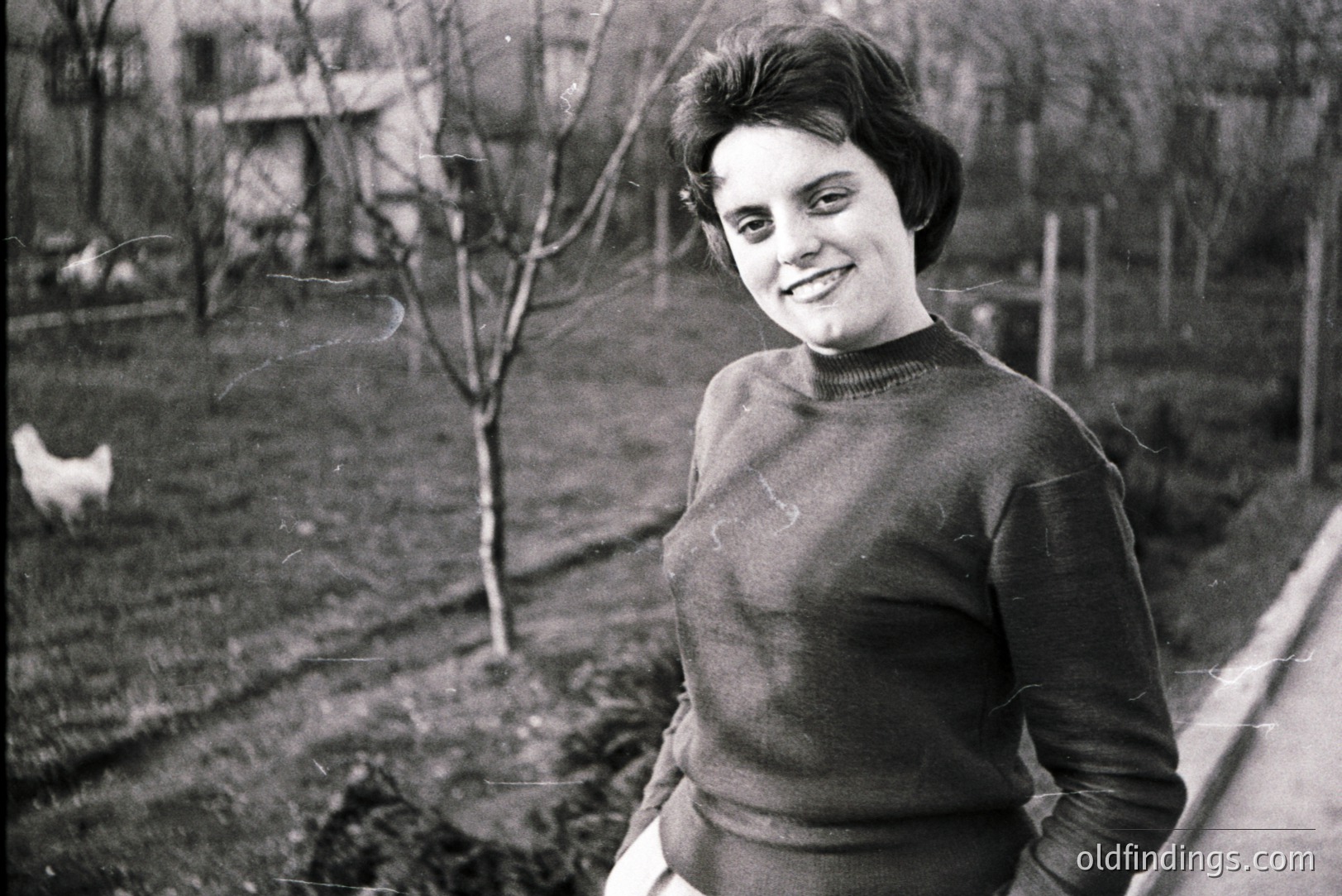 A smiling woman with a short, dark, bouffant hairstyle stands outdoors in a rural setting. She wears a dark, ribbed sweater and light-colored trousers. A chicken is visible in the background, alongside a glimpse of a building and a narrow road. Likely 1960s, Eastern European origin. Potential stock value for vintage fashion or lifestyle references.