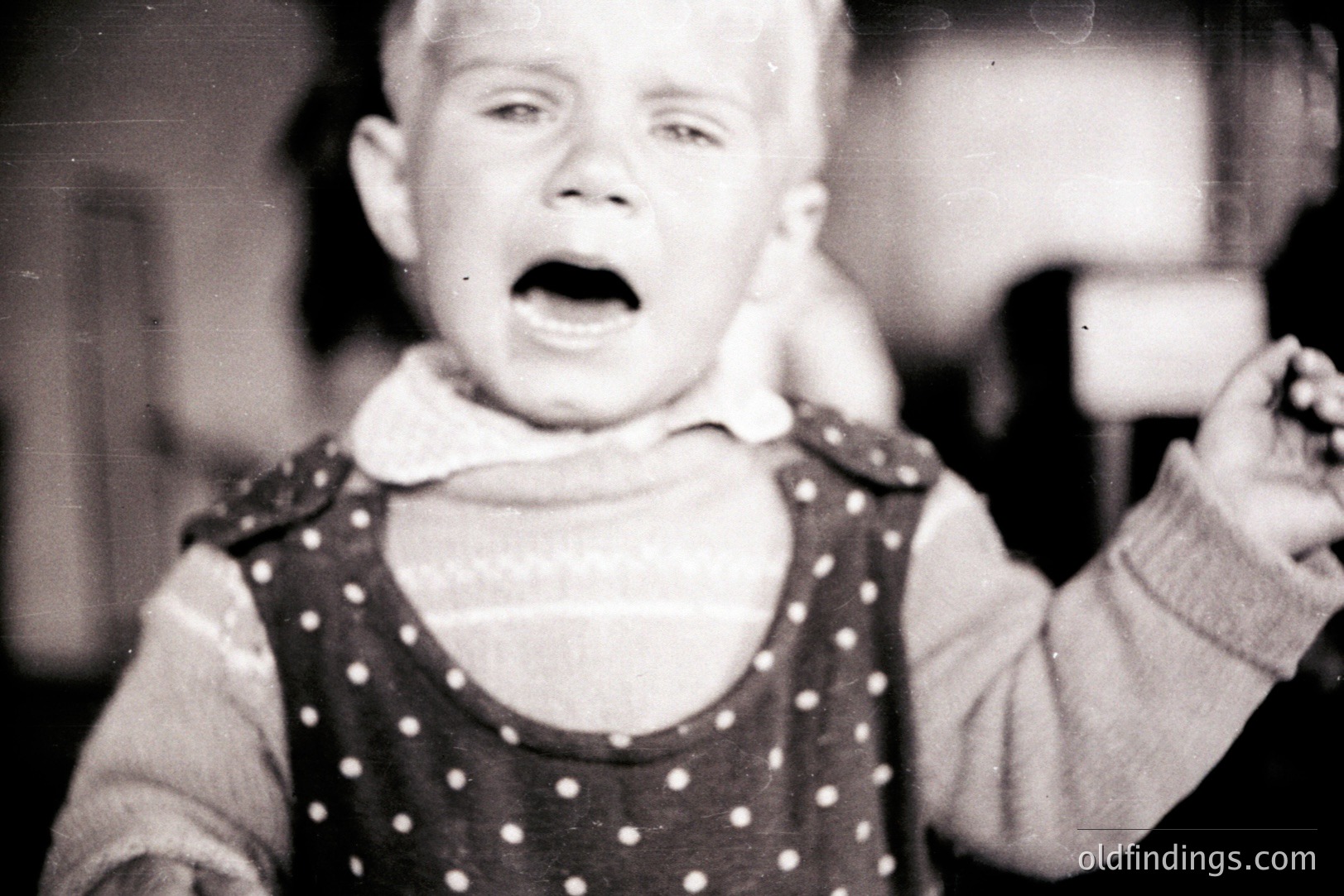 Close-up, black and white portrait of a young child crying with an open mouth. The child wears a patterned dress over a sweater. Likely a candid snapshot, possibly 1950s-1970s. The shallow depth of field creates a blurred background, highlighting the subject's distress.