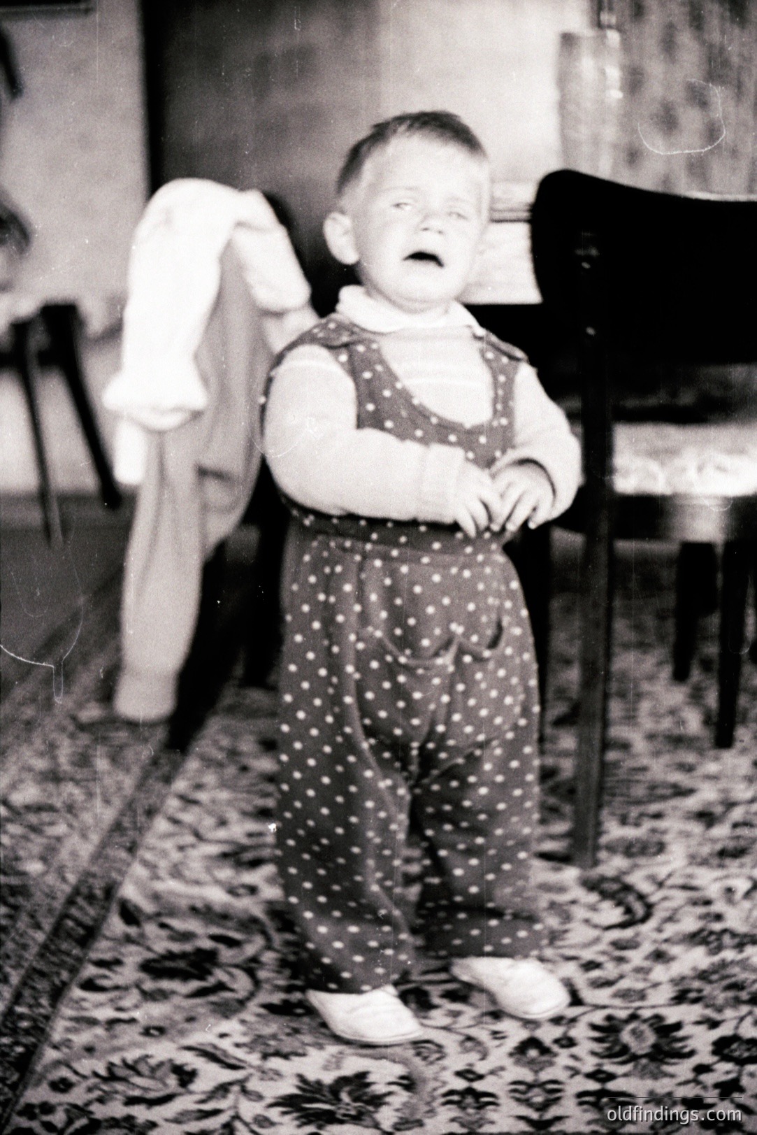 A young child, seemingly distressed, stands on an ornate rug, wearing a polka-dot romper and small shoes. The background includes furniture and a patterned wall, suggesting an interior setting. Likely a family snapshot from the early to mid-20th century. A poignant, candid moment.