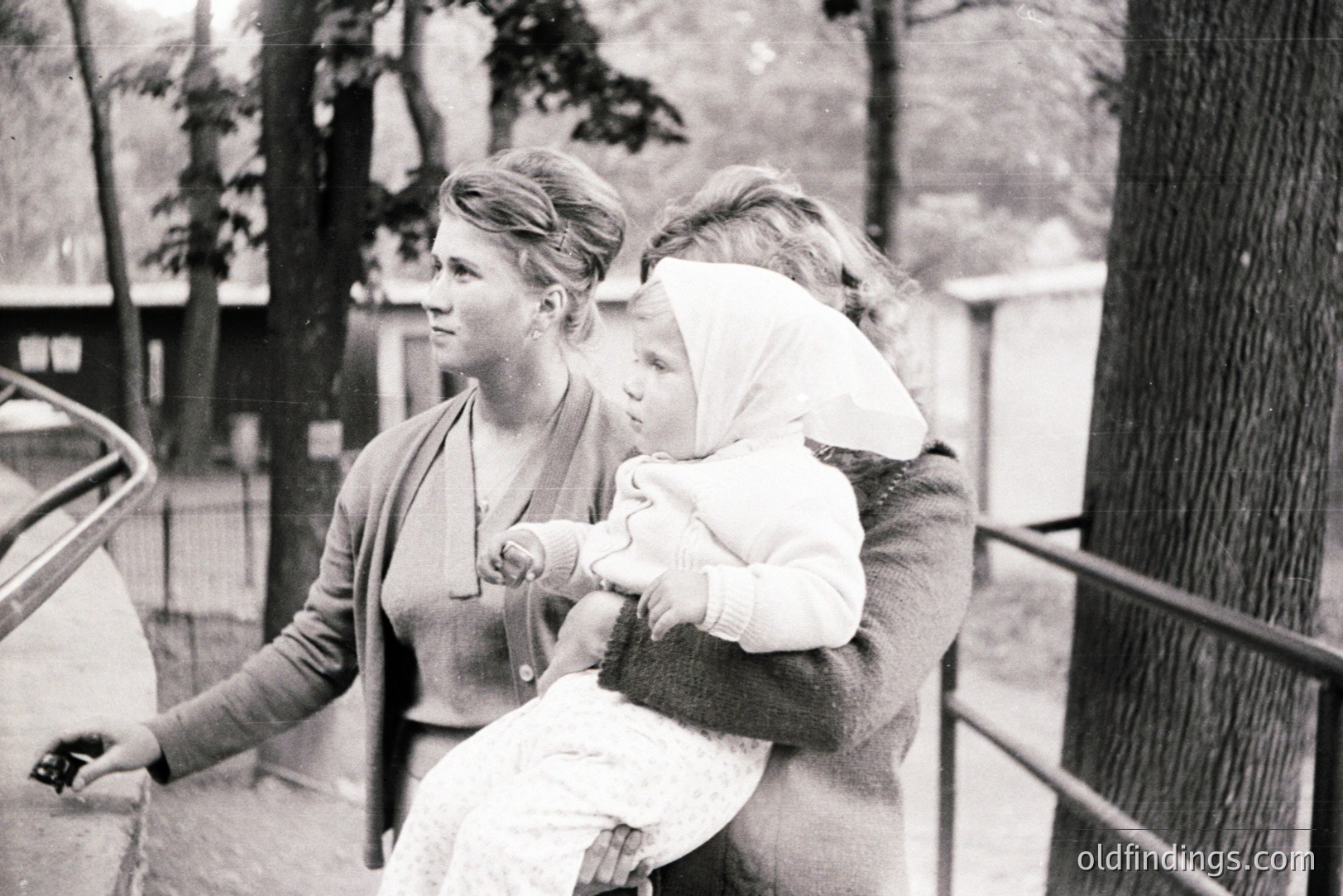 A woman in a cardigan holds a child bundled in a white bonnet, standing outdoors near a railing. The scene suggests a public park or garden setting, with trees visible in the background. Likely a candid moment, likely 1960s-1970s.