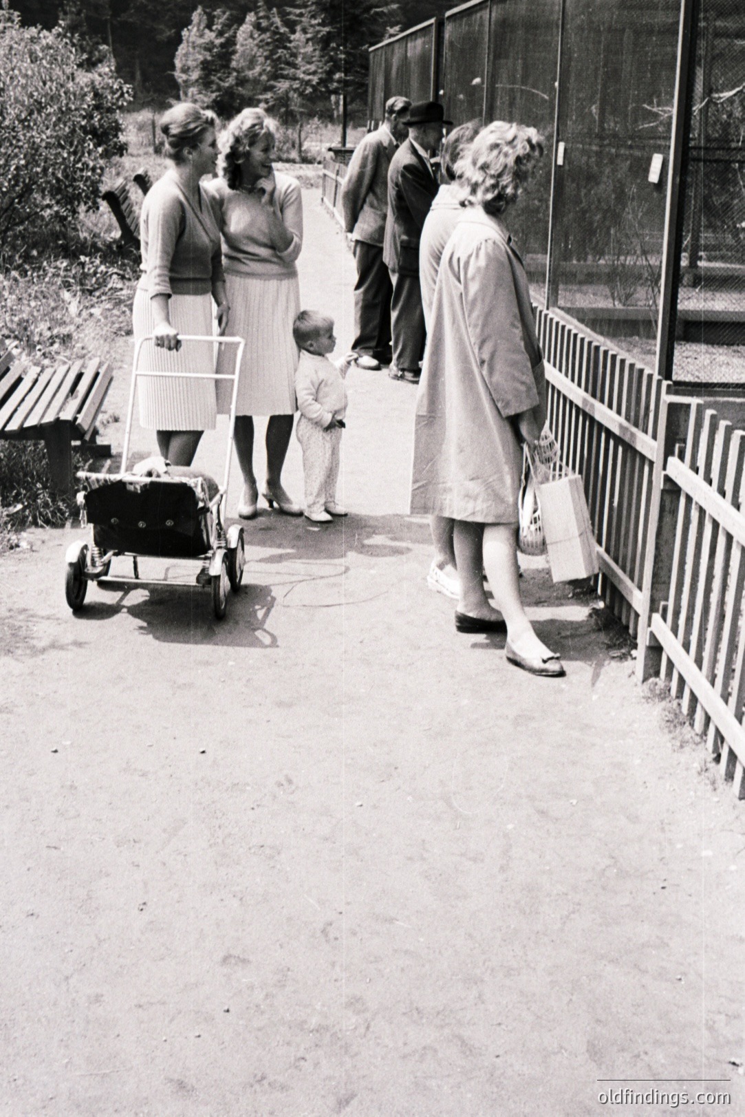 A family observes animals at a zoo, likely in the 1960s. A toddler in shorts gazes intently through a fence while adults in mid-century dresses and suits look on. A vintage stroller sits nearby. Demonstrates leisure activities and family dynamics of the era.