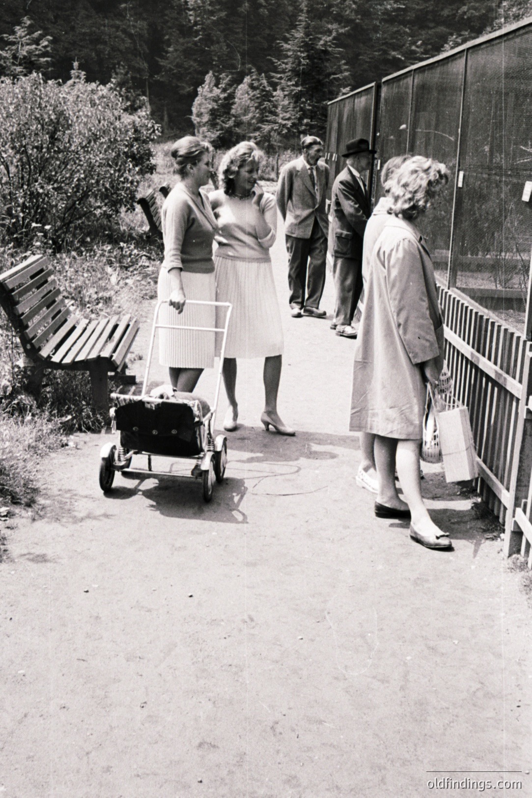 A group of four individuals—two women, two men—walk along a paved path near a fenced enclosure. The women wear 1960s-era dresses; one pushes a vintage wheeled pram. The men are in suits and a hat. Possibly a zoo or park setting. Likely mid-1960s.